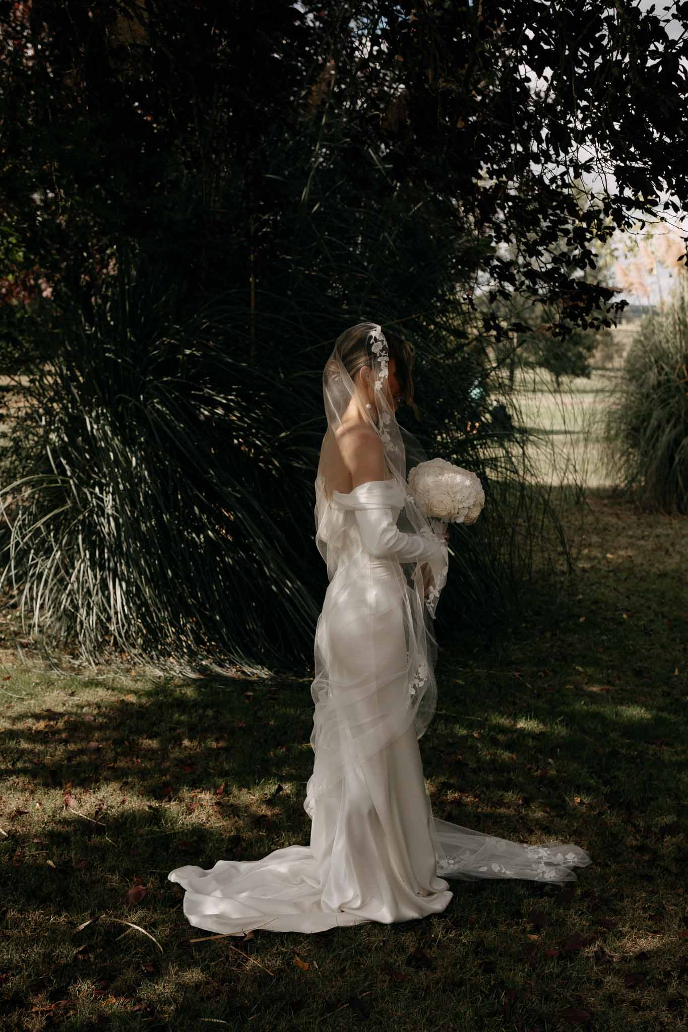 Bride from behind in fitted ivory satin gown with lace-edged veil and white peony bouquet in dappled garden light