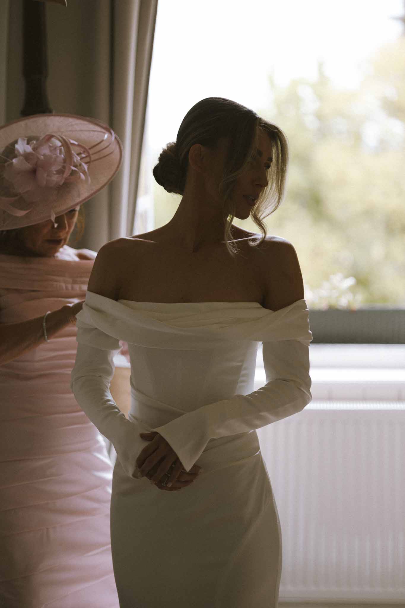 Mother in blush fascinator fastening bride's off-shoulder ivory gown at bright window