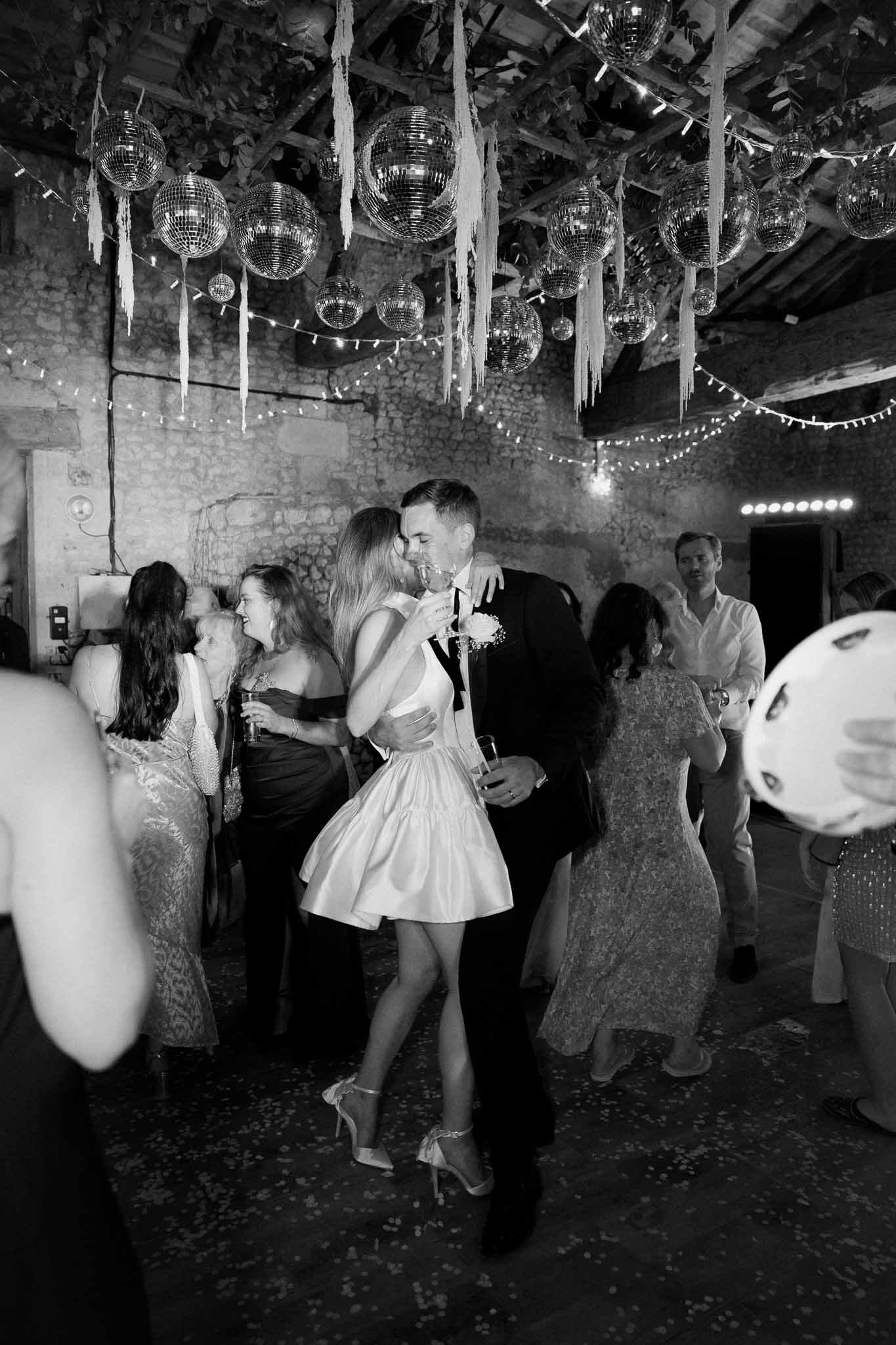 Black and white dance floor kiss beneath disco balls and fairy lights in rustic stone barn