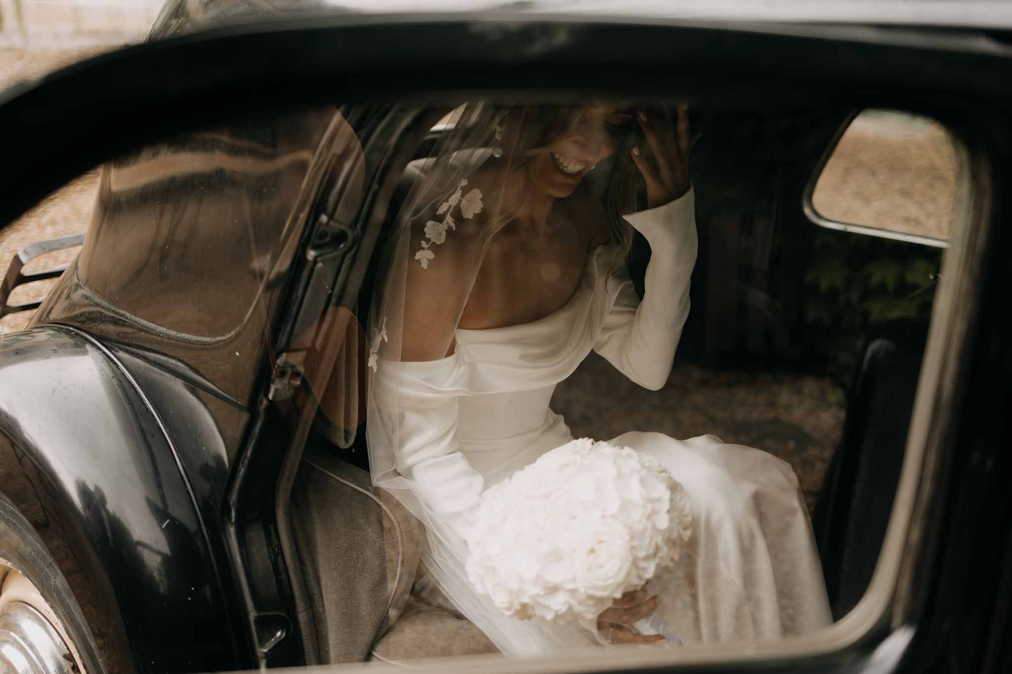 Laughing bride holding white peony bouquet seated in vintage black car wearing veil with floral applique