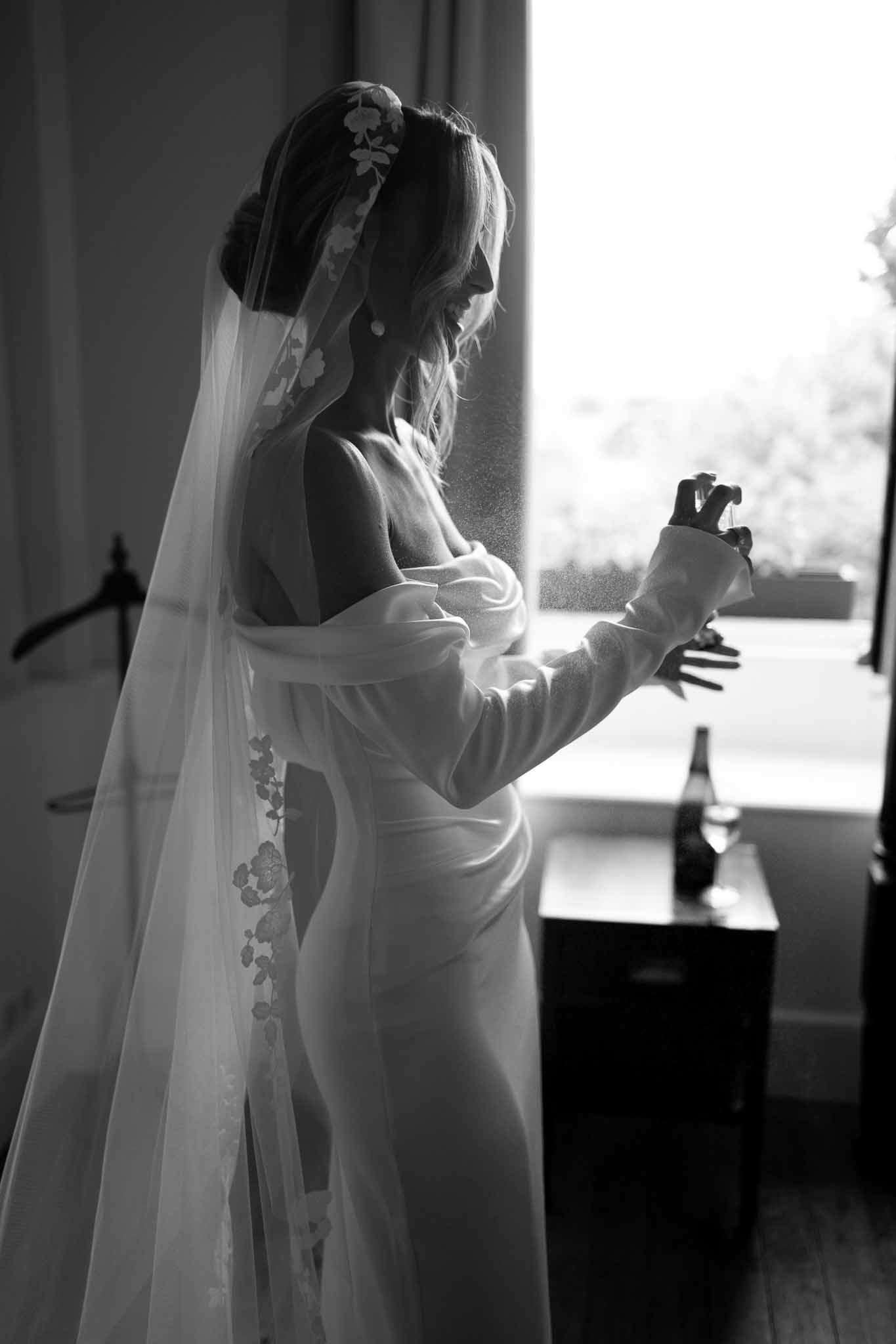 Black and white bride spritzing perfume by window with cathedral veil floral embroidery and champagne nearby