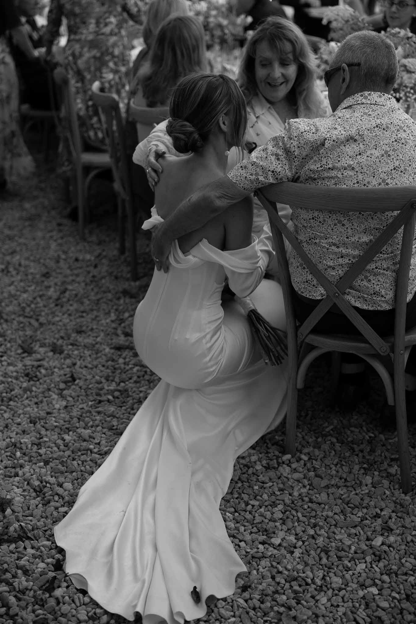 Bride crouches to embrace seated male guest at outdoor gravel reception with cross-back chairs in B&W