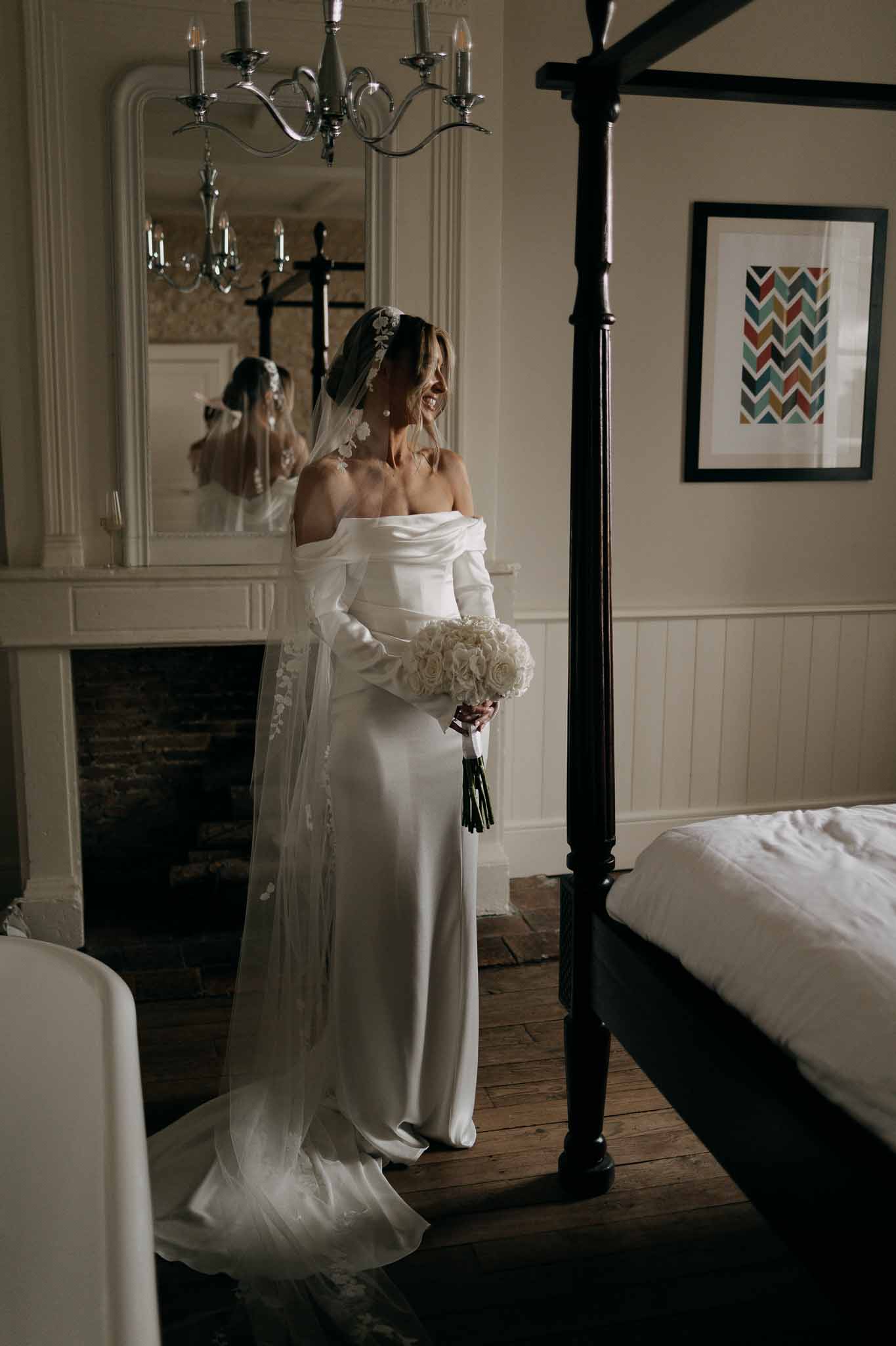 Bride in off-shoulder satin gown with floral applique veil holding white rose bouquet in four-poster bedroom