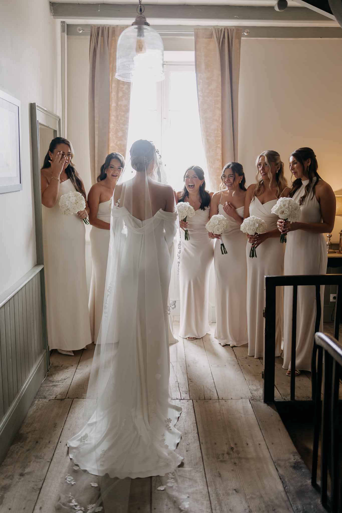 Bride reveals dress to six bridesmaids in ivory dresses holding white rose bouquets with emotional reactions