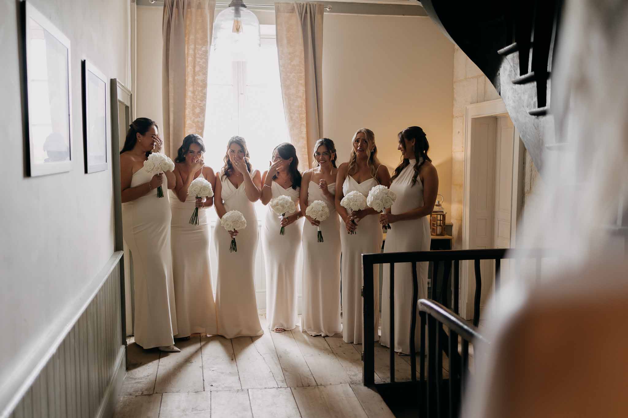 Six bridesmaids in ivory satin slip dresses reacting emotionally on chateau upper landing