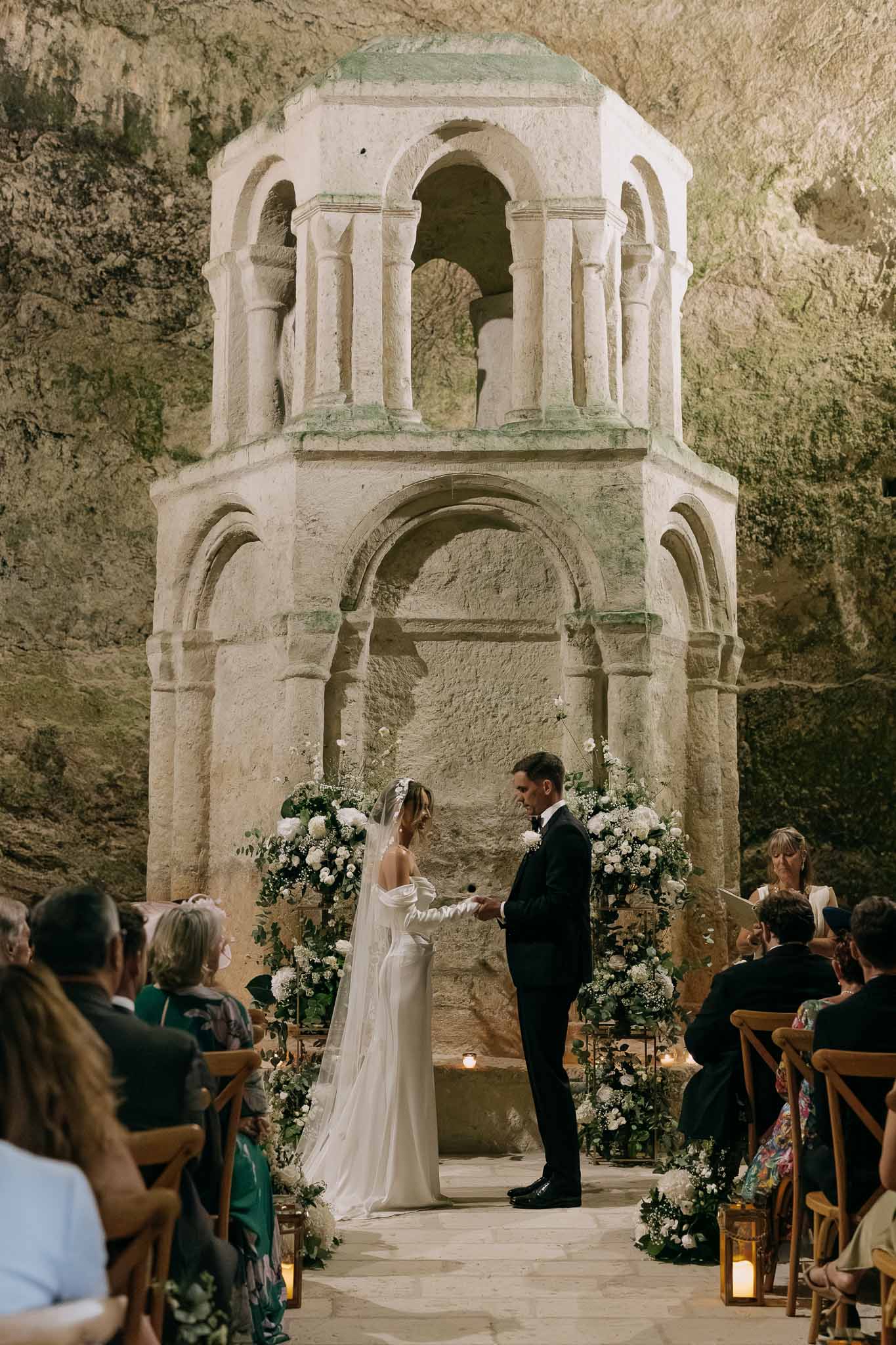 Wedding ceremony inside underground grotto with Romanesque stone altar, white and green floral arrangements, and gold lant...