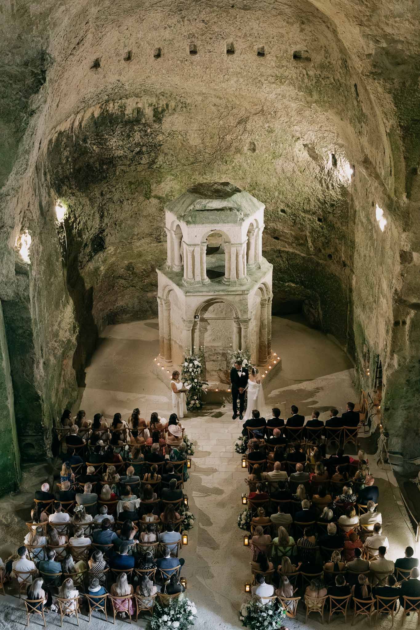 Wedding ceremony inside rock-hewn cave chapel with stone altar, white florals, and 100 guests in crossback chairs