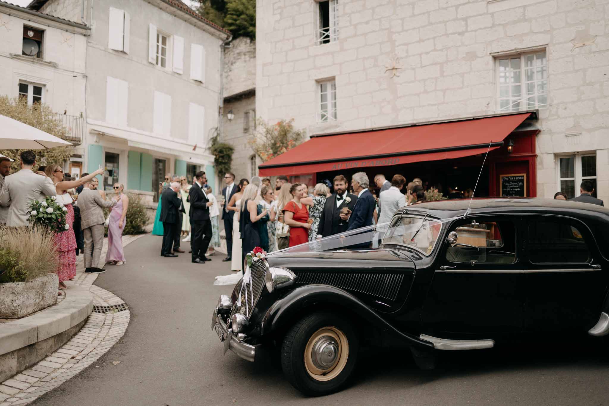Guests gathering around vintage black Citroen in French village square during cocktail hour