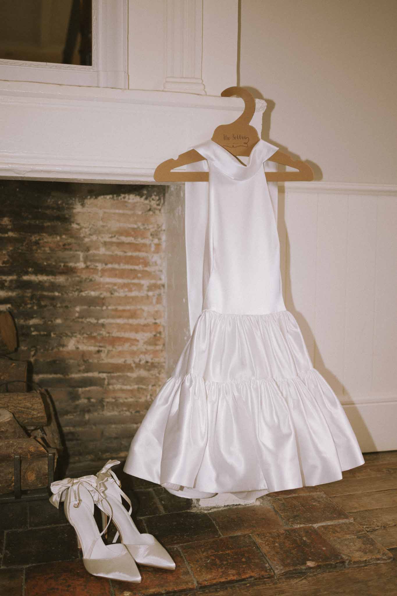 White satin halter mini dress on branded hanger with bow-tied pointed heels on brick hearth against panelled wall