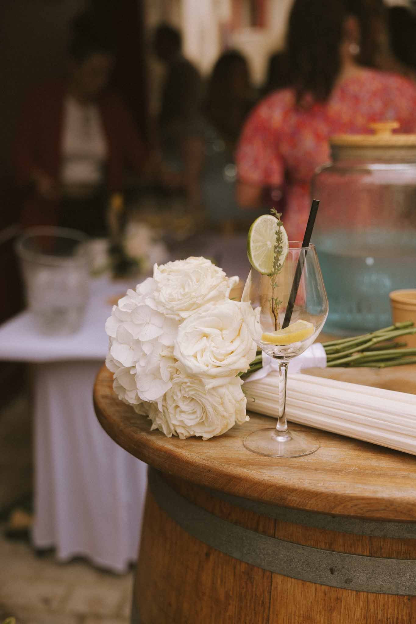 Bridal bouquet of ivory roses and white hydrangeas on wine barrel beside cocktail glass with lime garnish