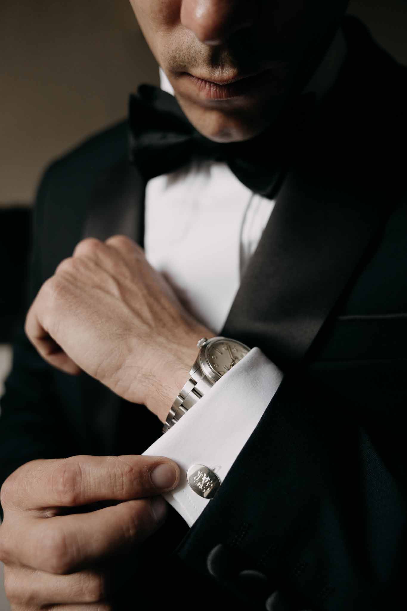 Groom fastening silver cufflink on white French cuff in black tuxedo with bow tie and silver watch