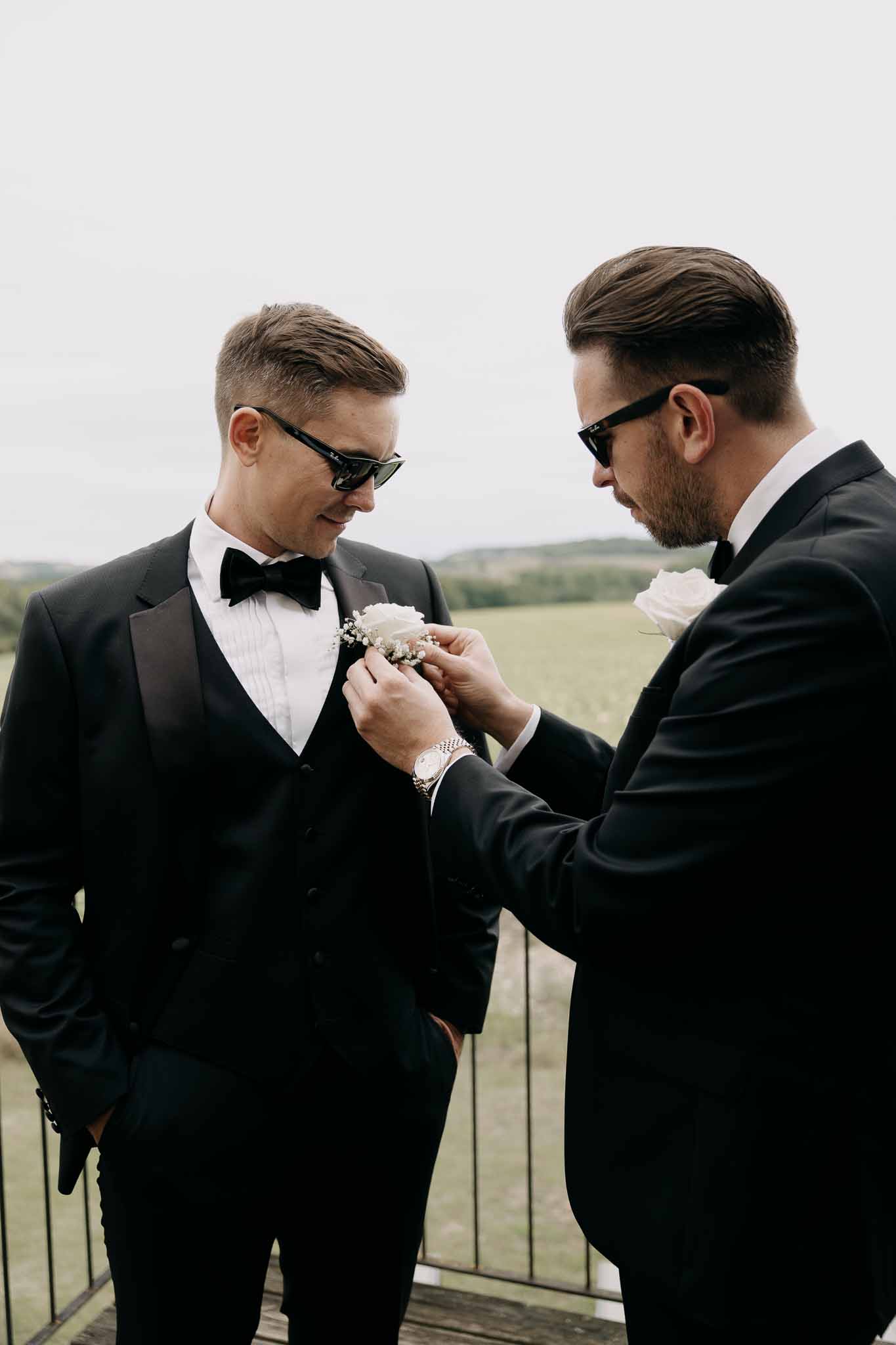 Groomsman pinning white rose boutonniere on groom's black tuxedo lapel on outdoor terrace