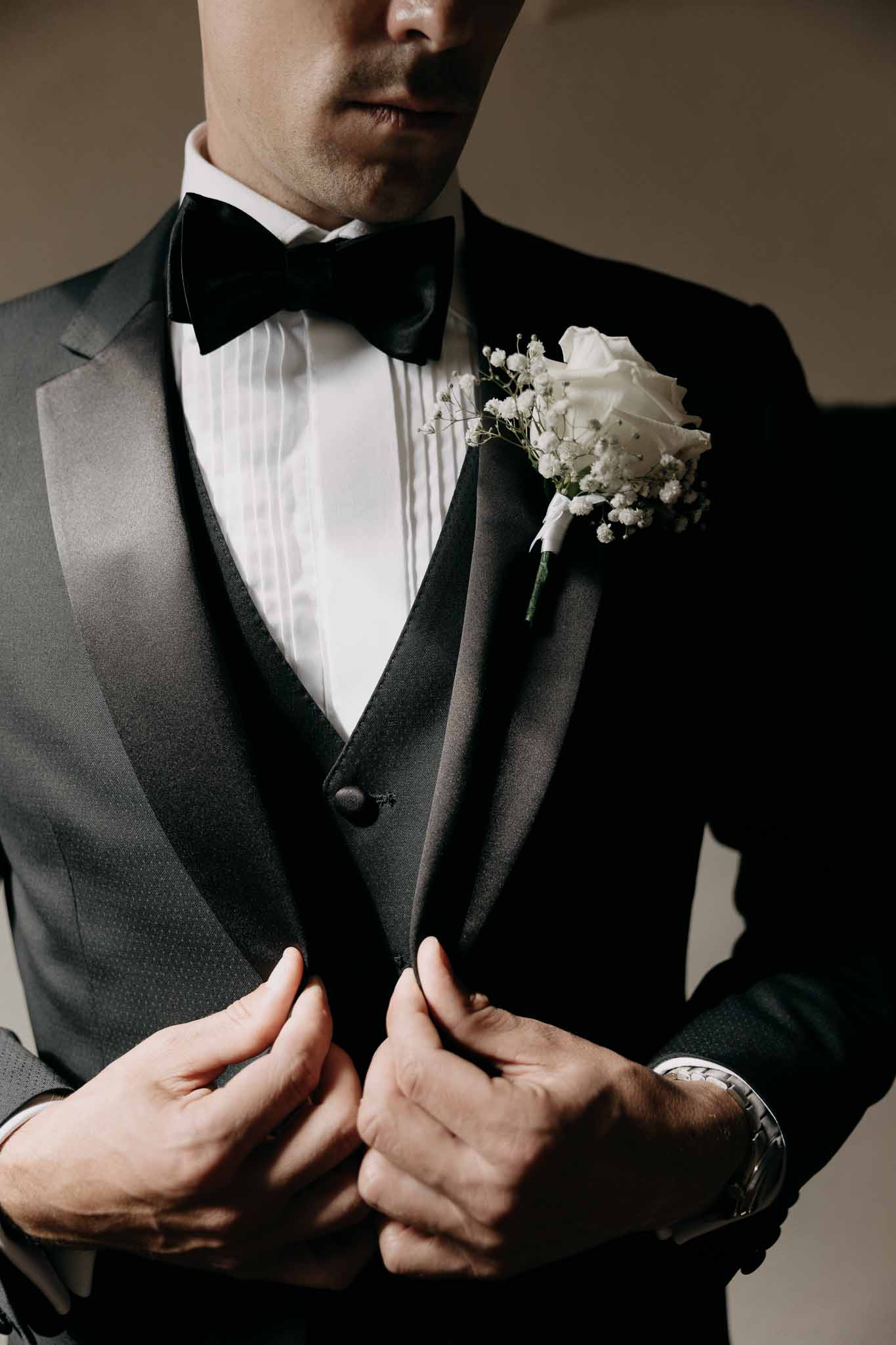 Groom adjusting charcoal tuxedo lapels with satin peak detail, black velvet bow tie, and white rose boutonniere