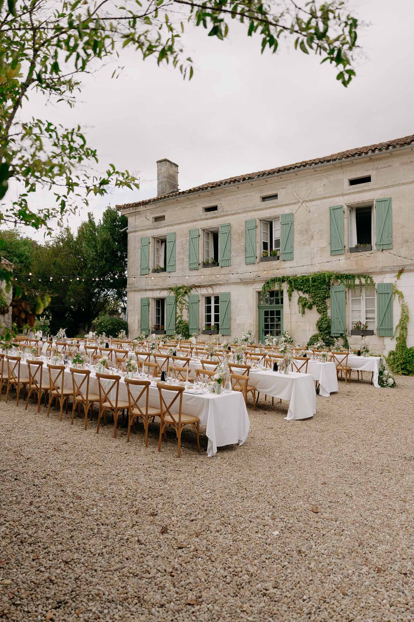 Outdoor reception tables with white linen and cross-back chairs in a French manor courtyard with fairy lights