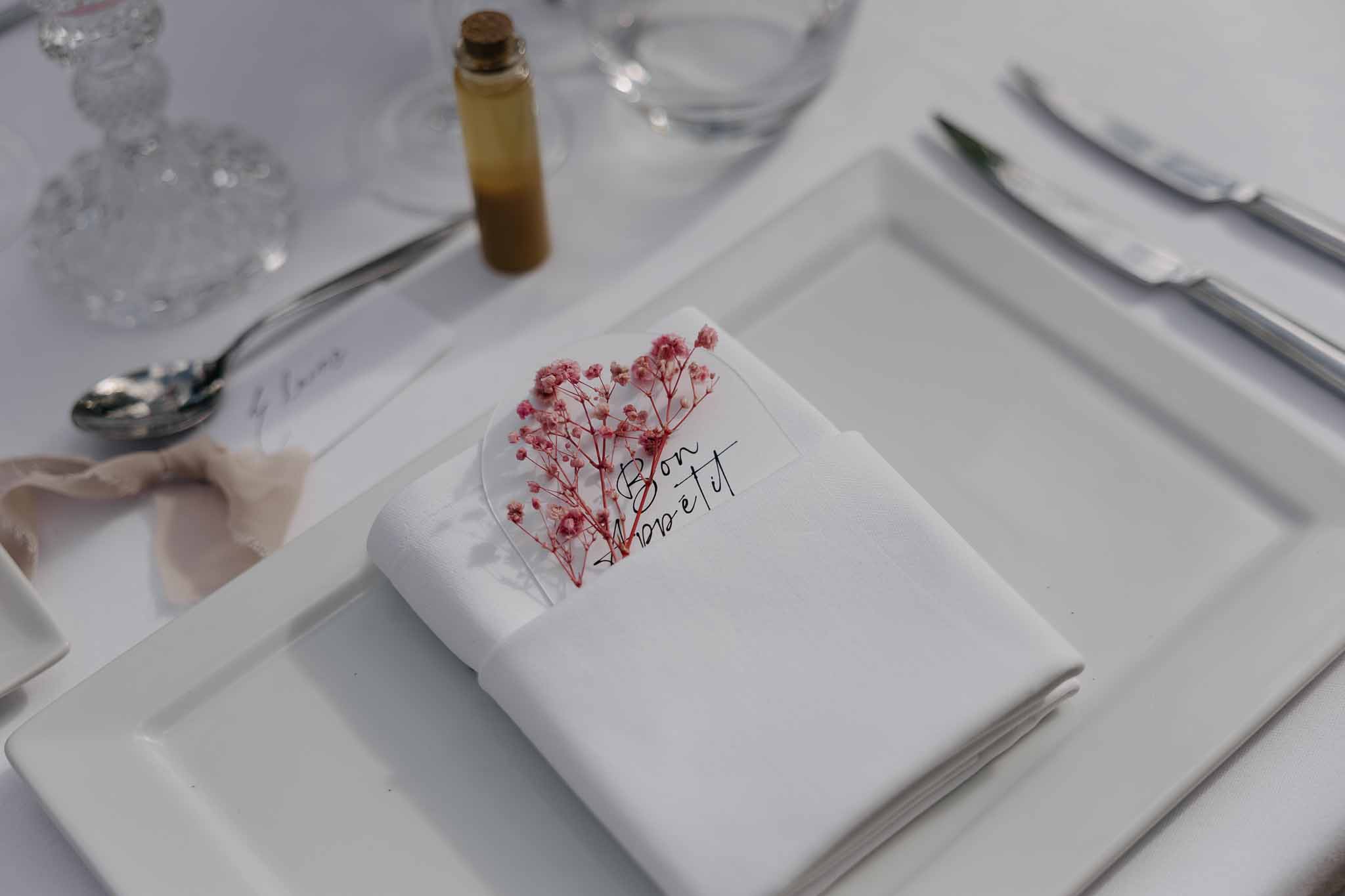 Place setting with heart-shaped acrylic tag dried pink gypsophila Bon Appetit text corked favor bottle on white linen