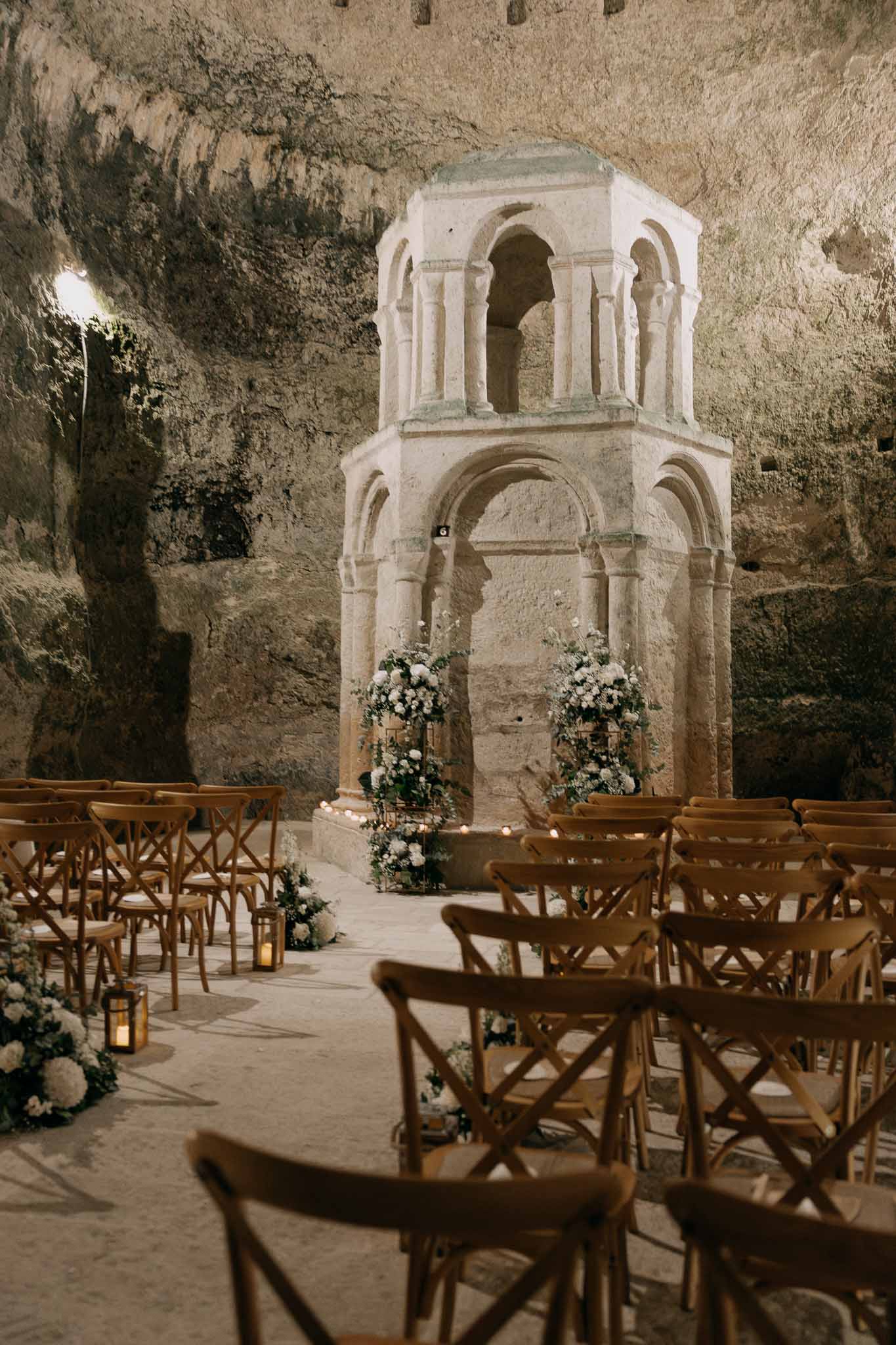 Romanesque stone ciborium altar flanked by white floral stands and candlelit aisle in vaulted troglodyte cave