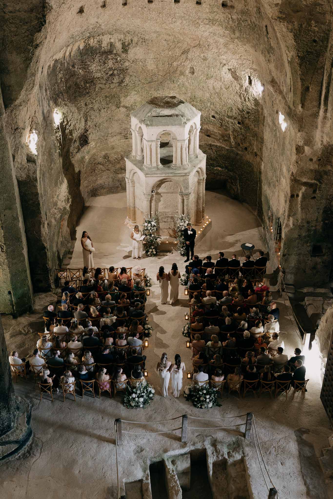 Couple at altar in troglodyte cave chapel with stone baldachin as 90 guests sit by candlelit aisle