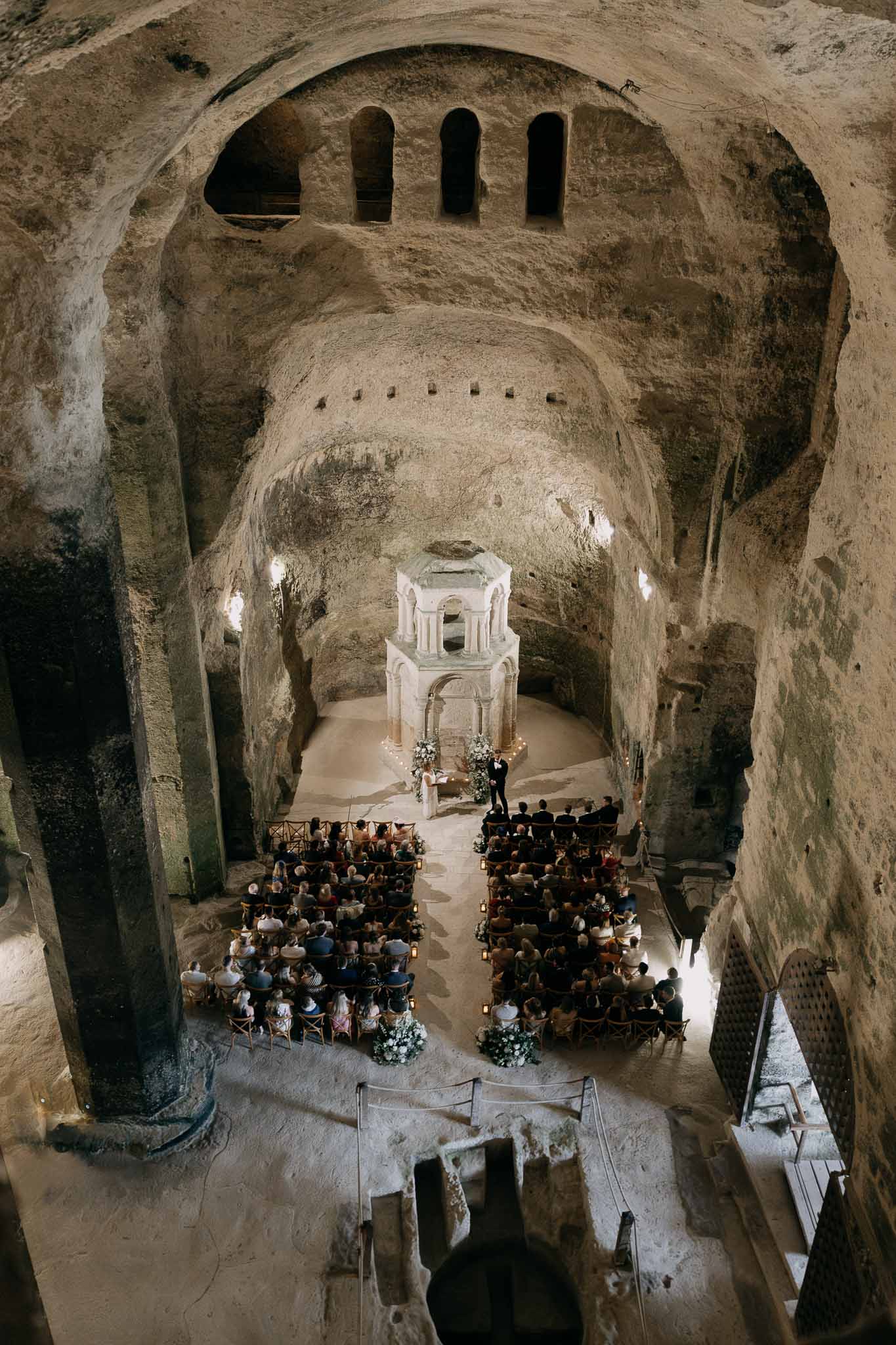 Aerial view of 100 guests in troglodyte chapel with lantern-lined aisle and white altar carved in limestone