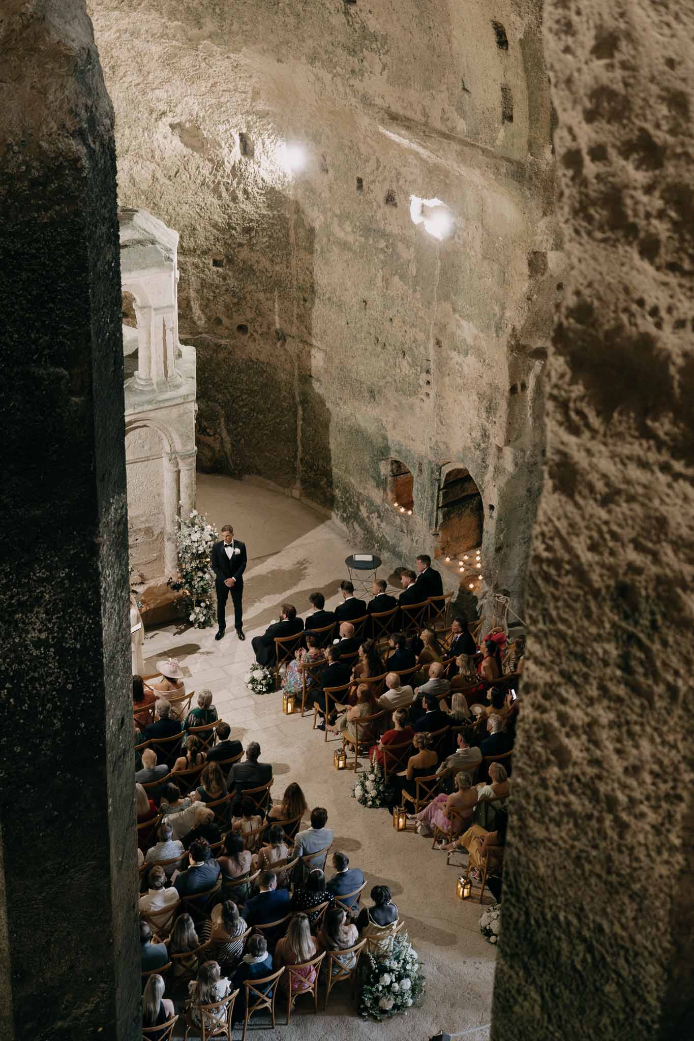 Wedding ceremony inside troglodyte cave chapel with cross-back chairs lanterns and white florals