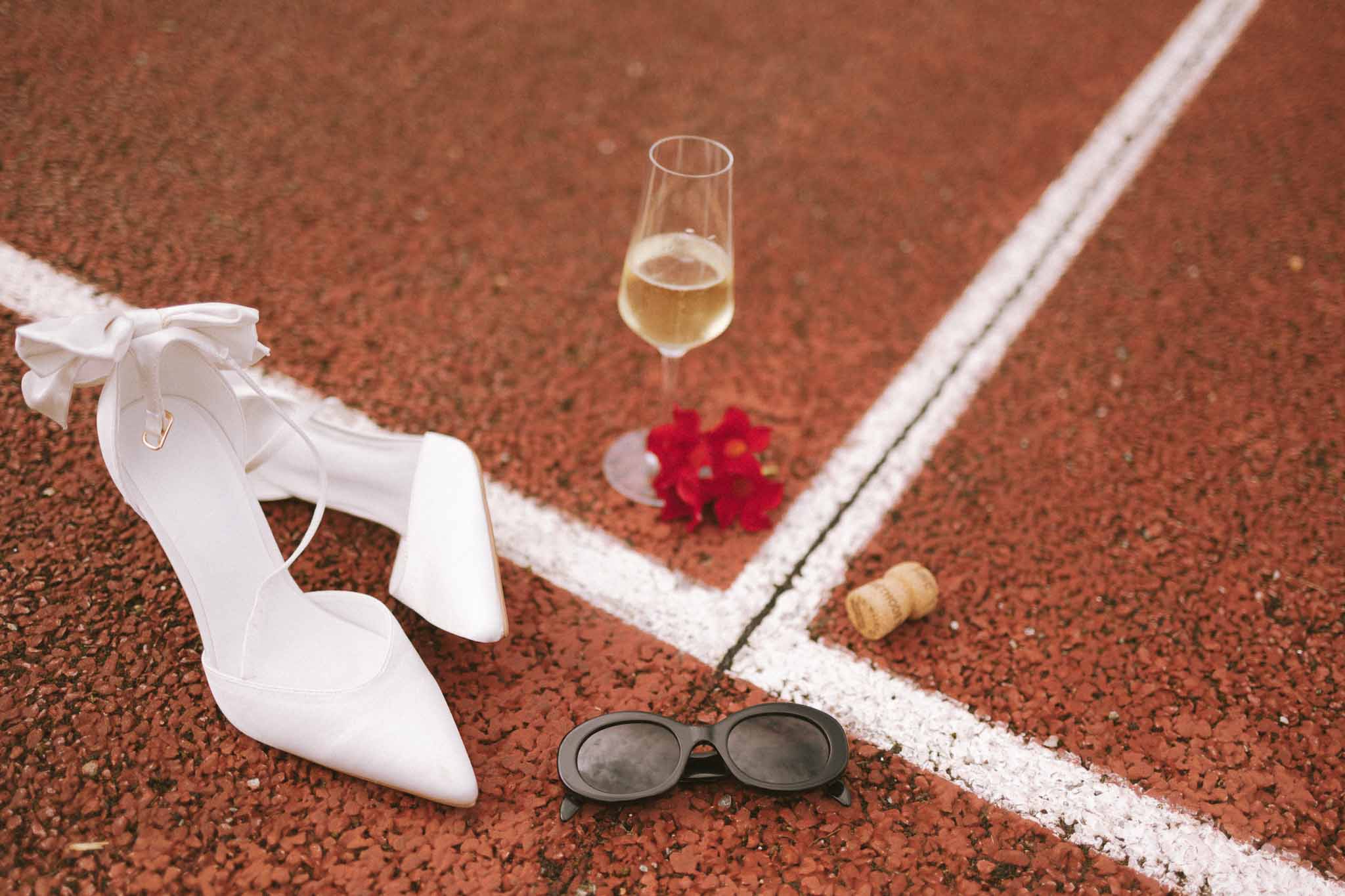 Bridal shoes, champagne flute, red flowers, and sunglasses arranged on a red clay tennis court