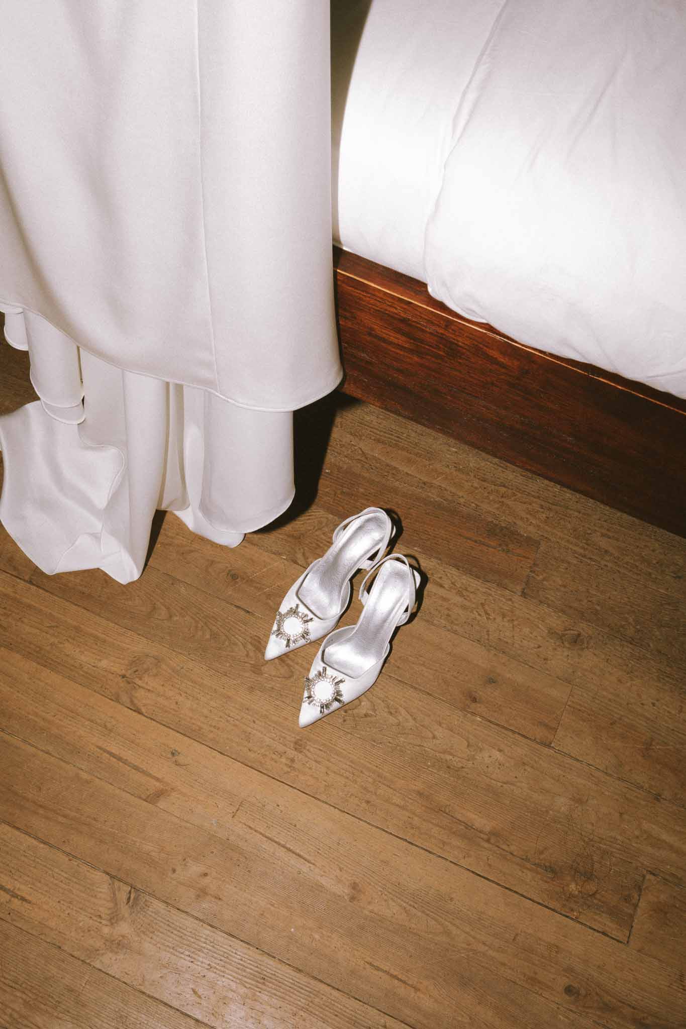 White satin slingback heels with crystal sunburst brooches beside ivory ruffle dress hem on hardwood floor