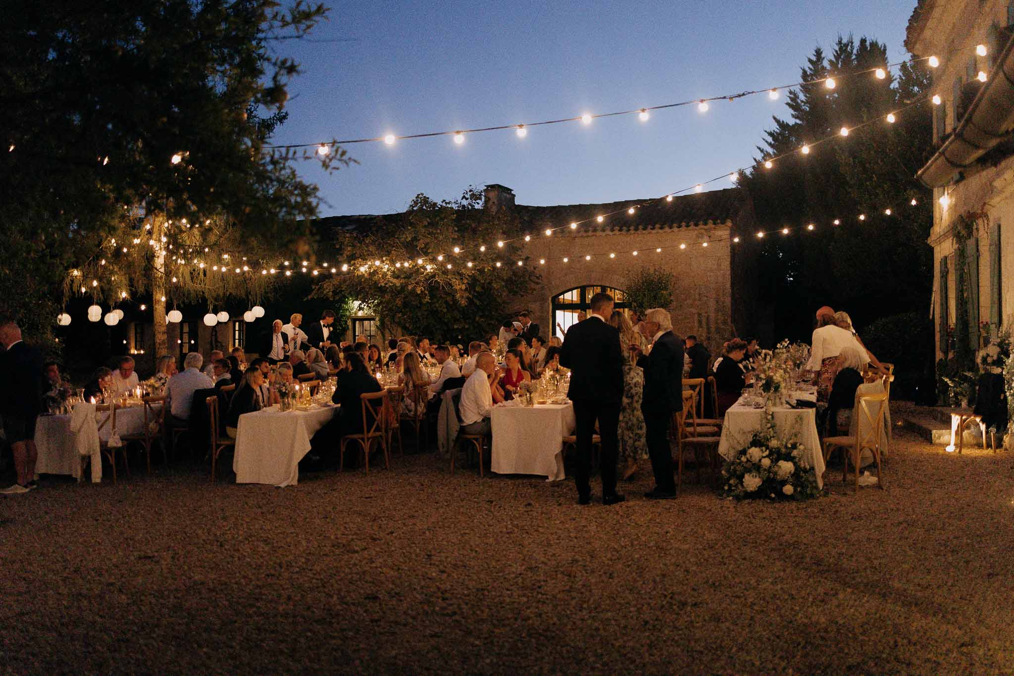 Outdoor courtyard reception at dusk with guests at long tables under festoon lights at French stone estate