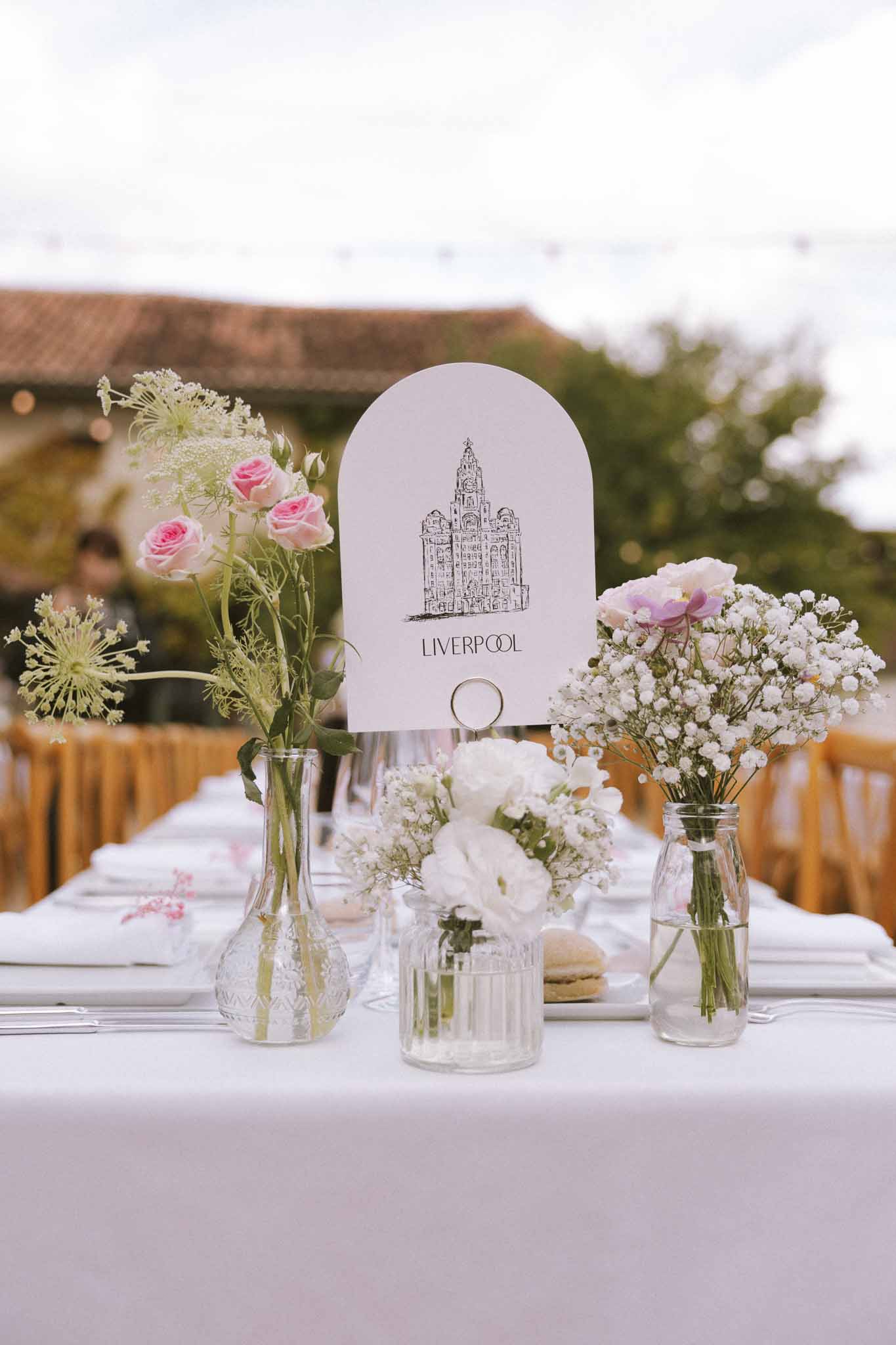 Mismatched glass bud vases with pink roses and queen anne's lace beside Liverpool table name card