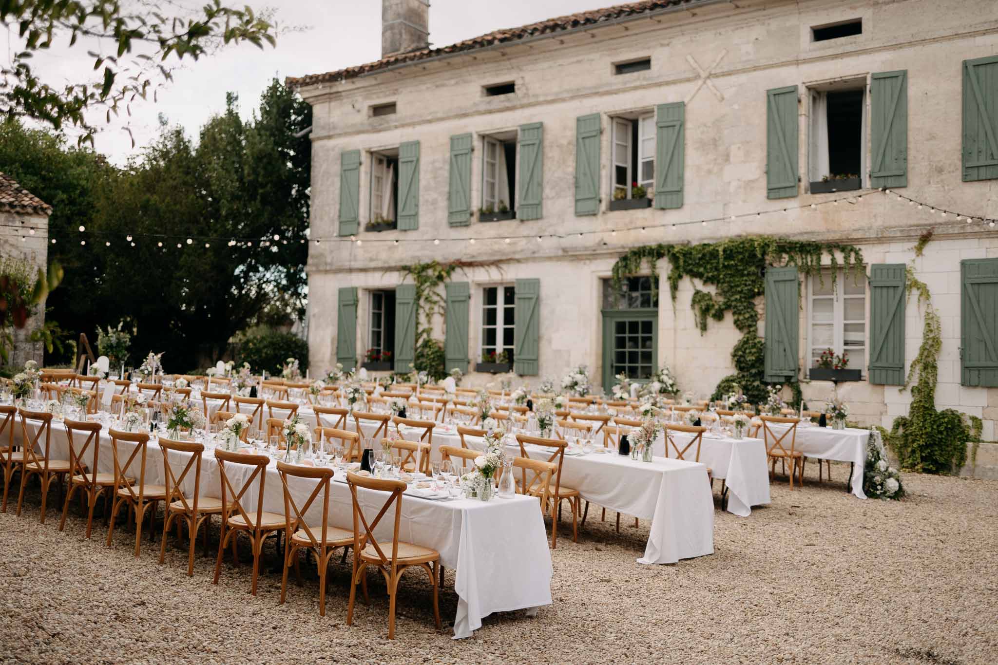 Banquet tables with wildflower bud vases and cross-back chairs under festoon lights at sage-shuttered manor