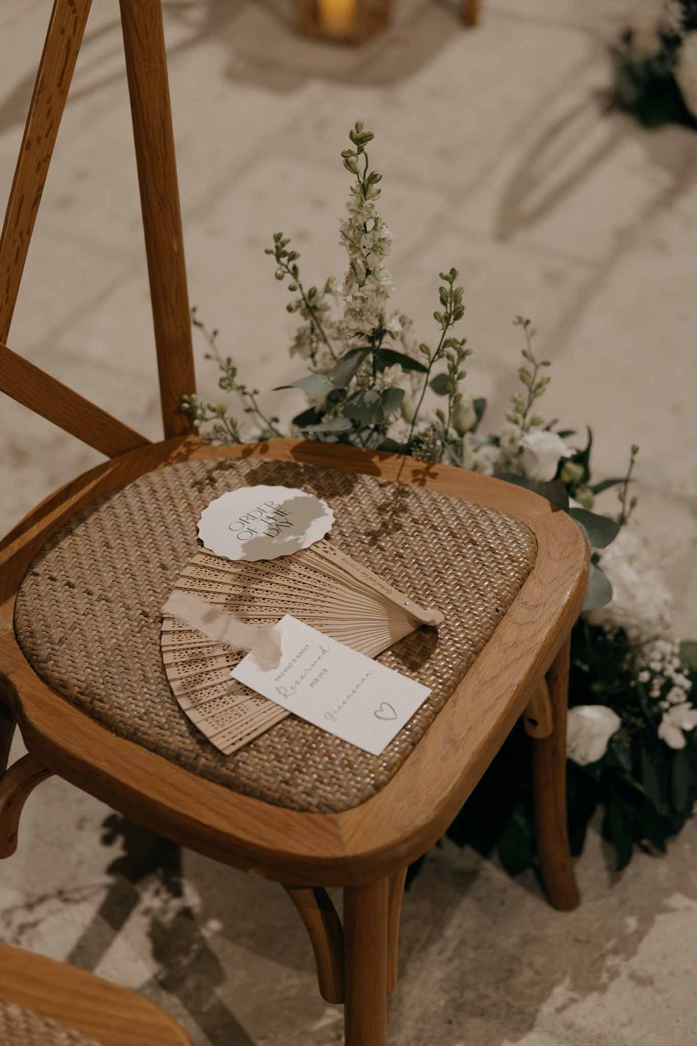 Wooden cross-back ceremony chair with hand fan order of day card and white floral arrangement at base