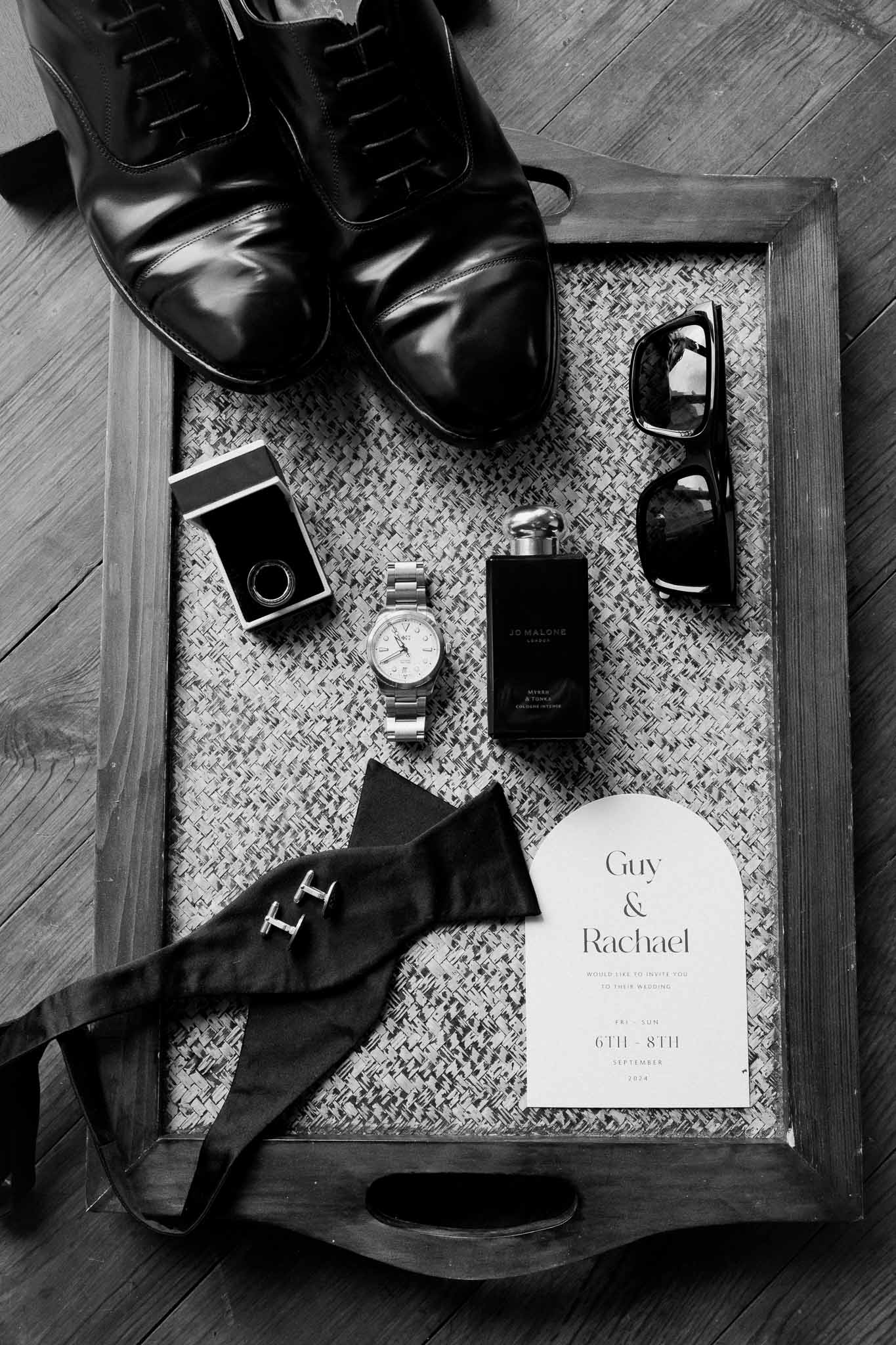 Groom flat lay with Oxford shoes, Jo Malone cologne, bow tie, ring box, and wedding invitation in B&W
