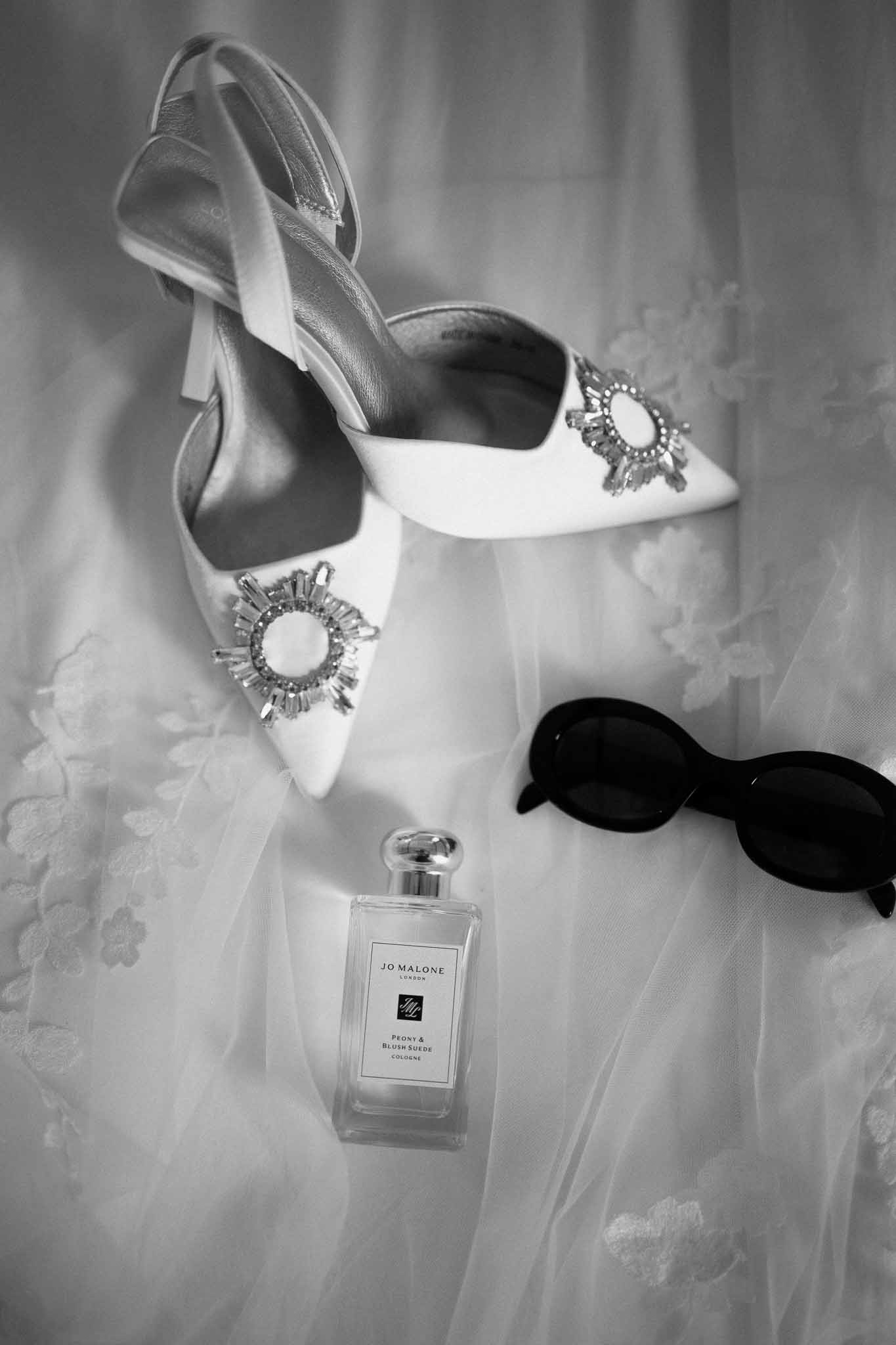 Black-and-white flat lay of crystal-embellished slingback heels, Jo Malone perfume, and sunglasses on lace veil