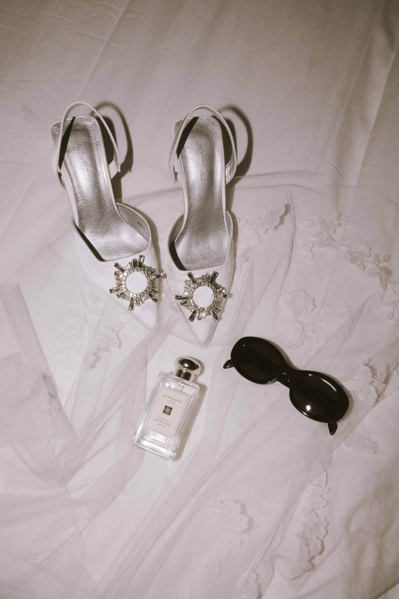 Flat lay of crystal-embellished slingback heels, Jo Malone cologne, and sunglasses on lace veil
