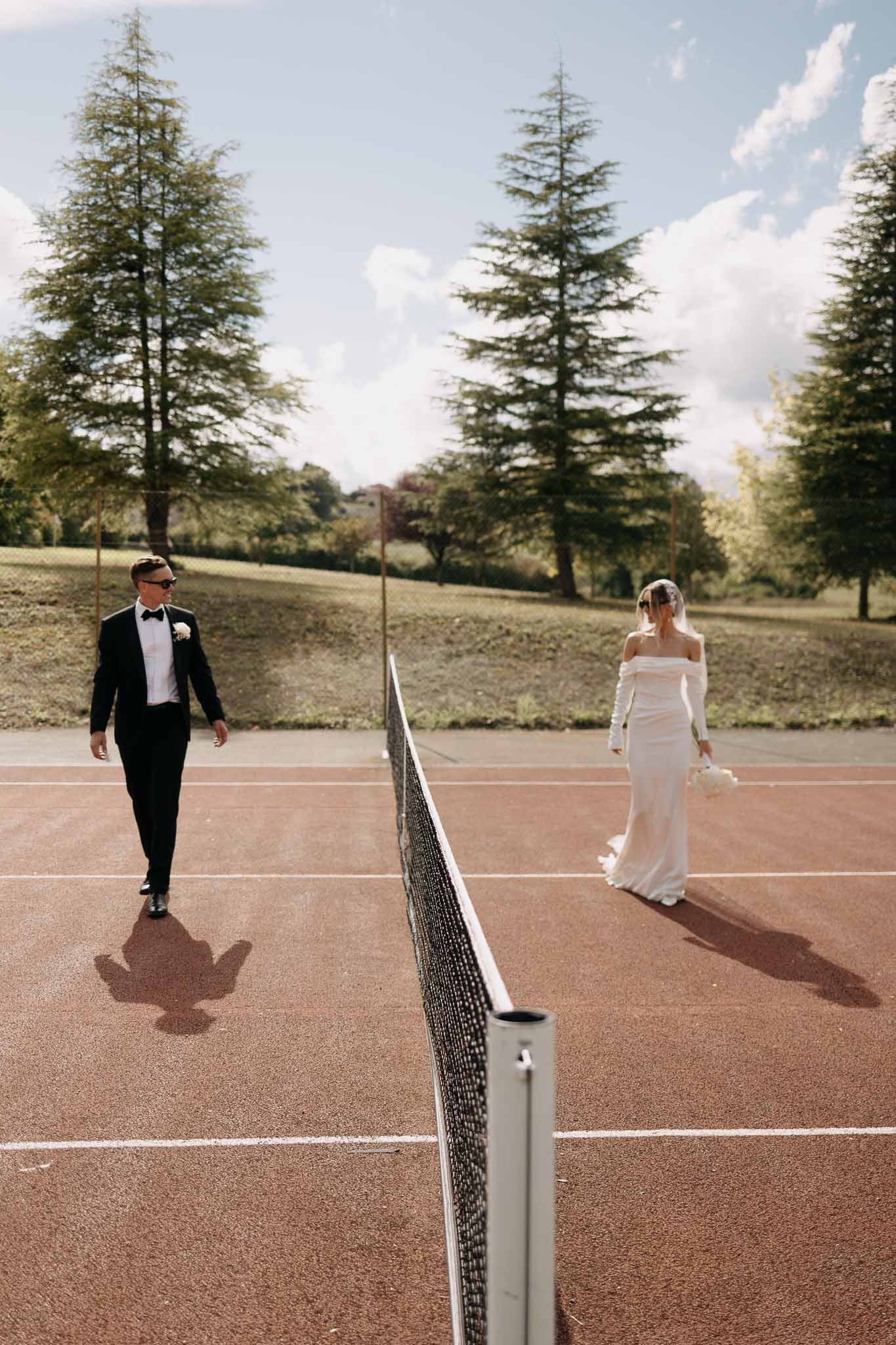 Couple walking on opposite sides of tennis court net, bride in off-shoulder gown with train and groom in tuxedo