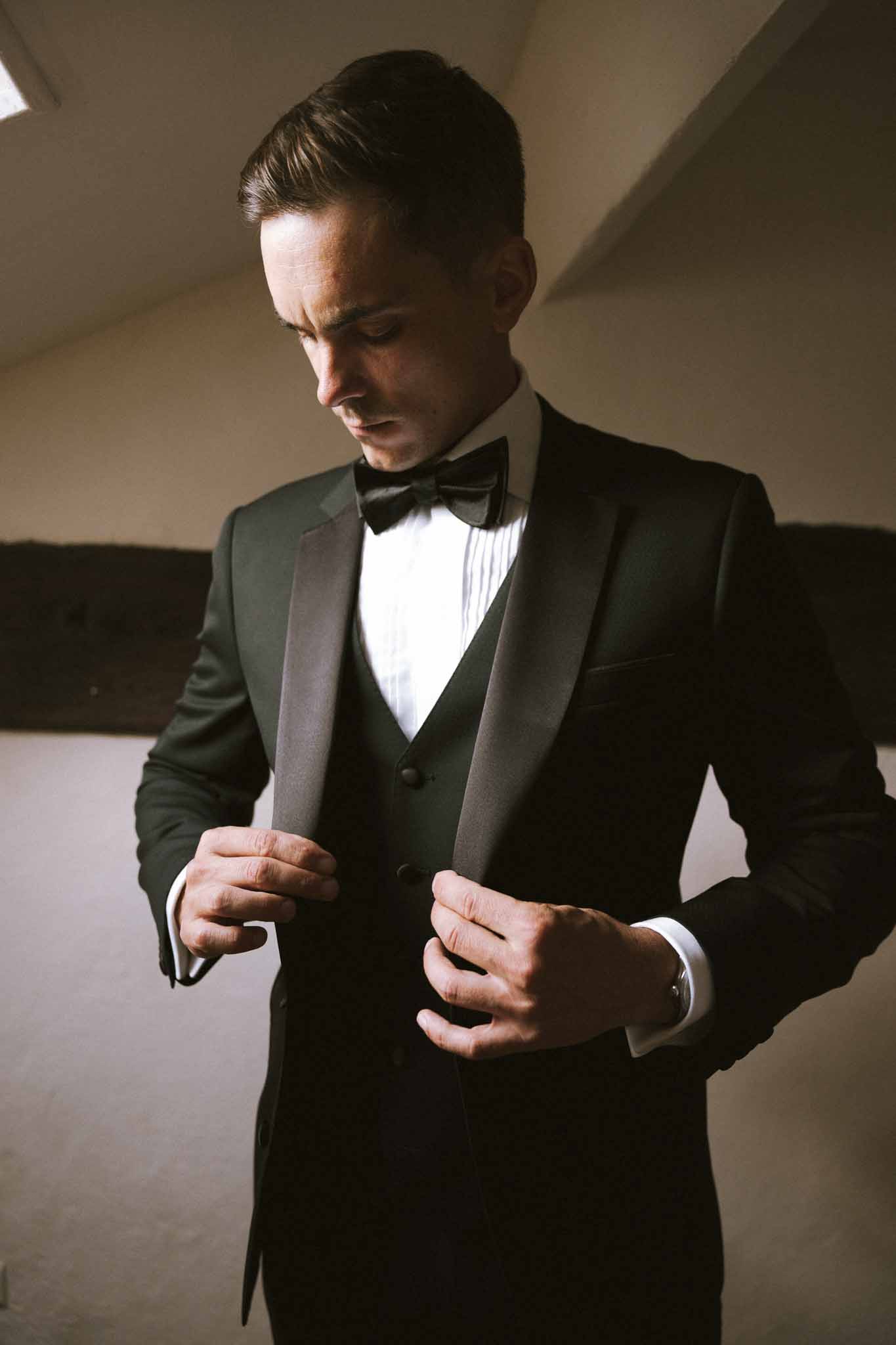 Groom adjusting tuxedo buttons with dramatic directional lighting during getting ready