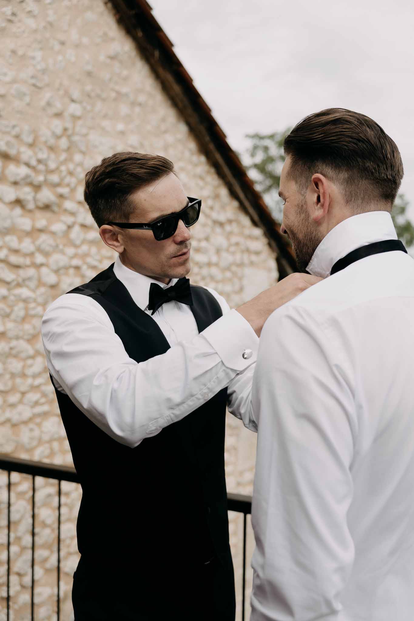 Groomsman in waistcoat and sunglasses adjusting another man's bow tie on stone building terrace