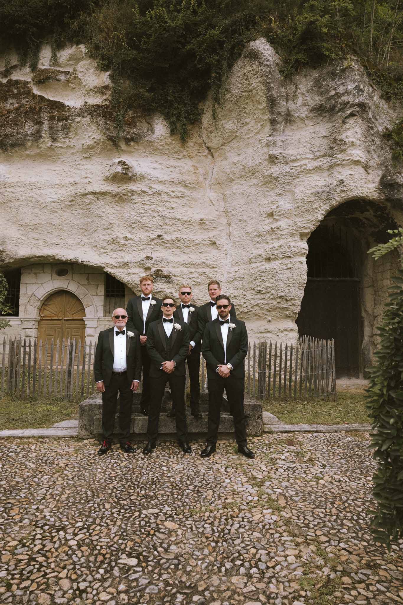 Six groomsmen in black tuxedos and sunglasses posed before carved tuffeau rock face on cobblestone