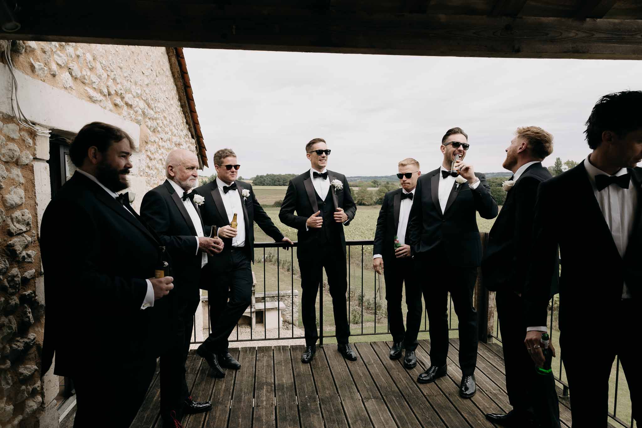 Groom and seven groomsmen in black tuxedos socializing on wooden terrace of stone farmhouse