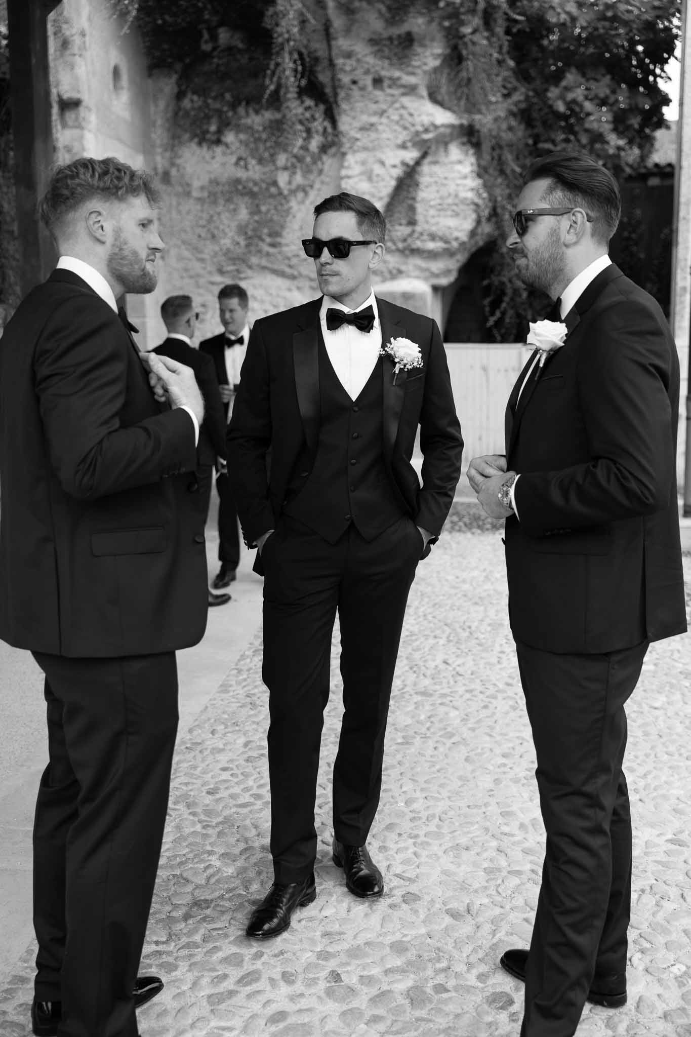 Groom in black tuxedo with sunglasses and two groomsmen converse on cobblestone before ceremony in B&W