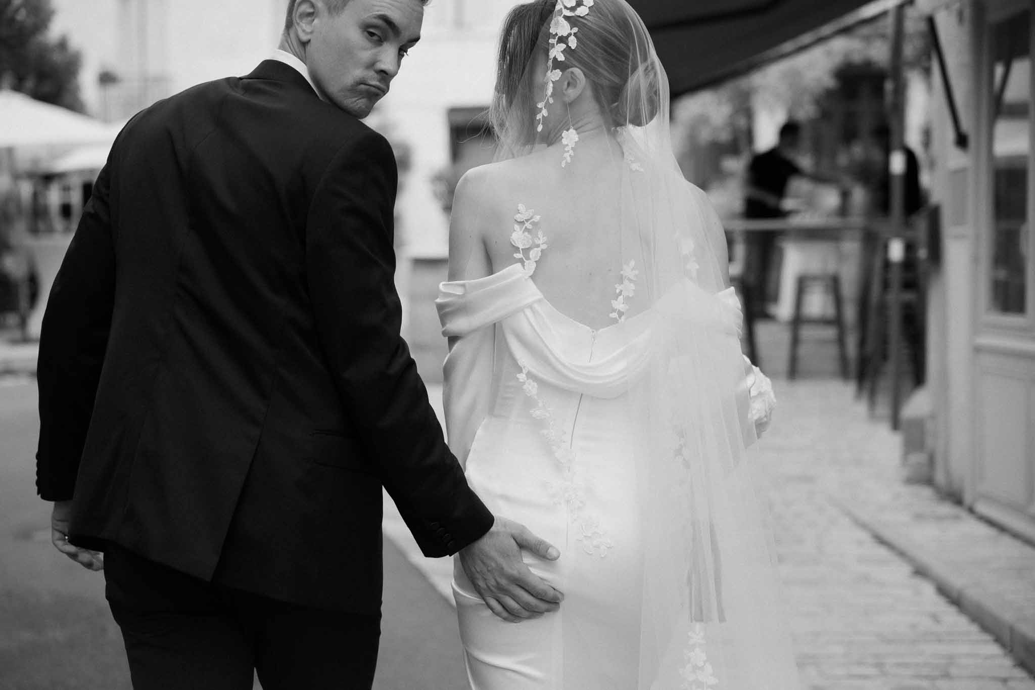 Black and white of couple walking on street from behind, bride in lace-back gown with floral-trimmed veil