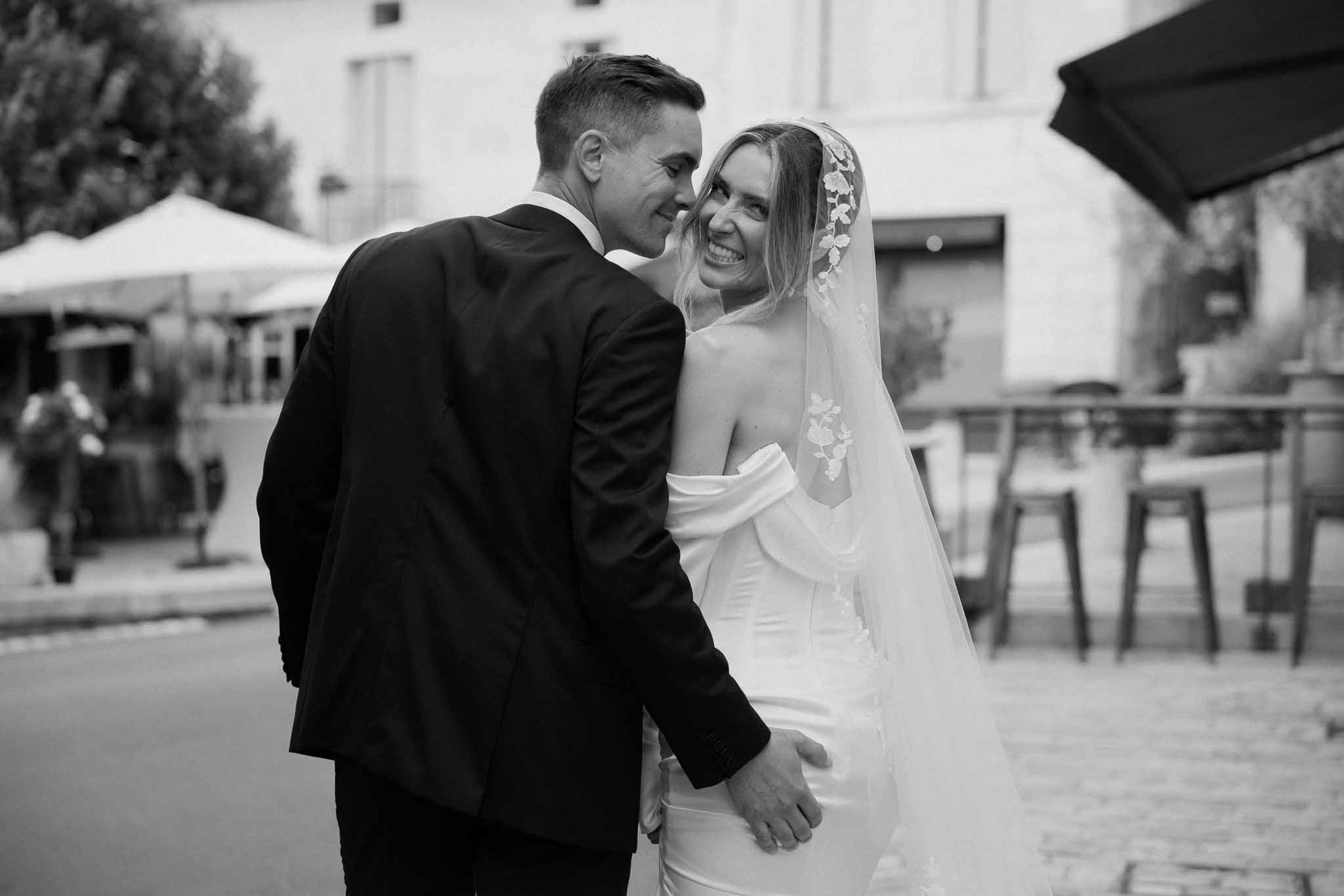 Groom embraces smiling bride in strapless gown with lace-edged veil on cobblestone street in B&W