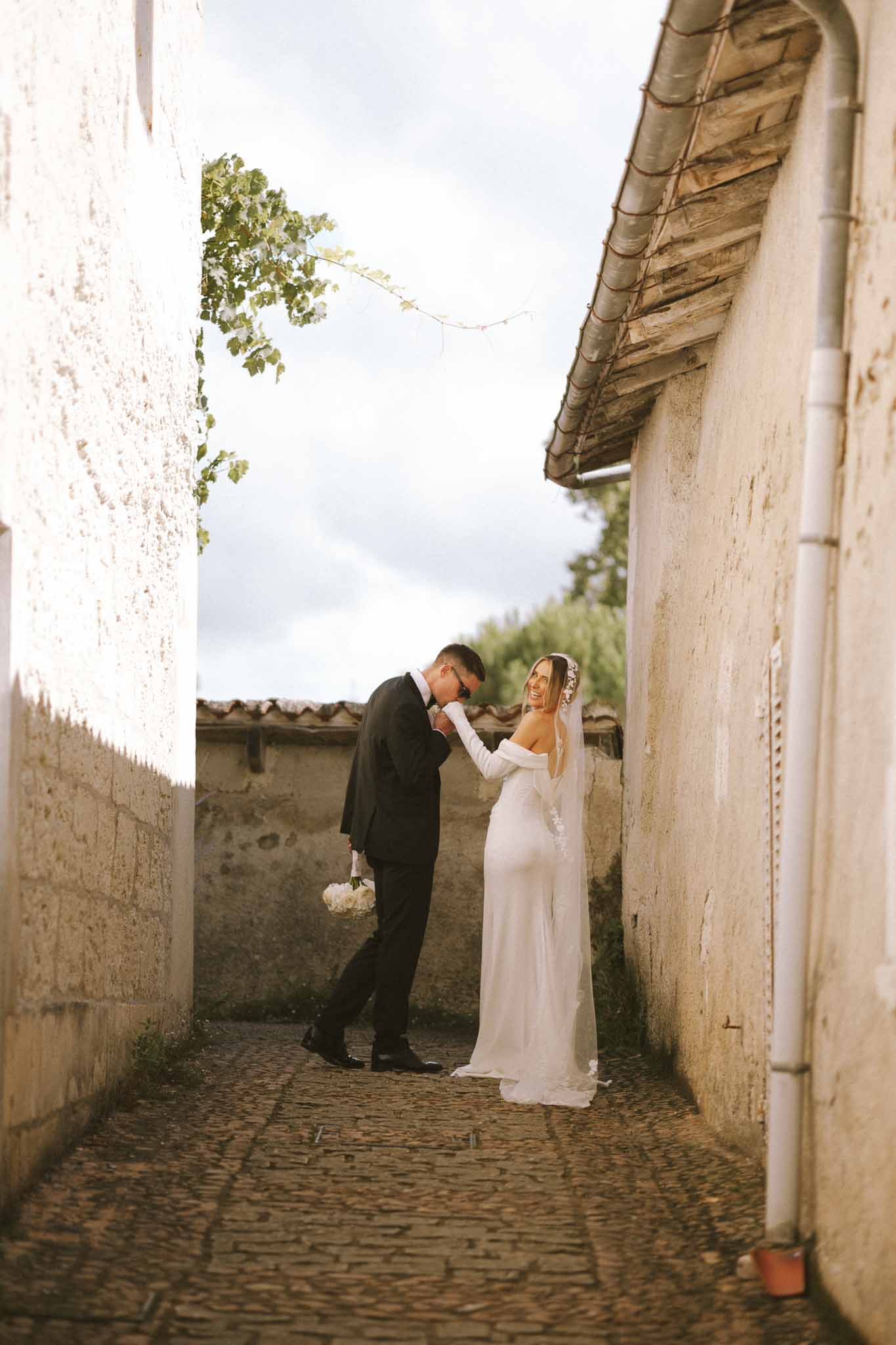 Groom in navy suit kissing bride's gloved hand in cobblestone passageway at French estate