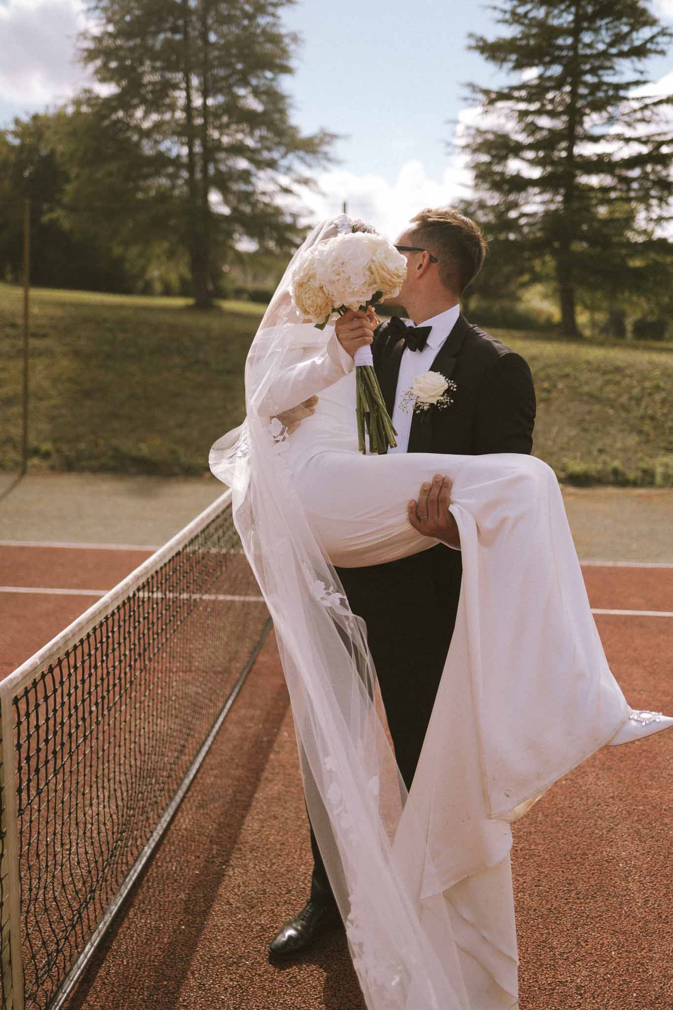 Groom lifts bride in his arms on a red clay tennis court as they kiss, bride holding white peony bouquet