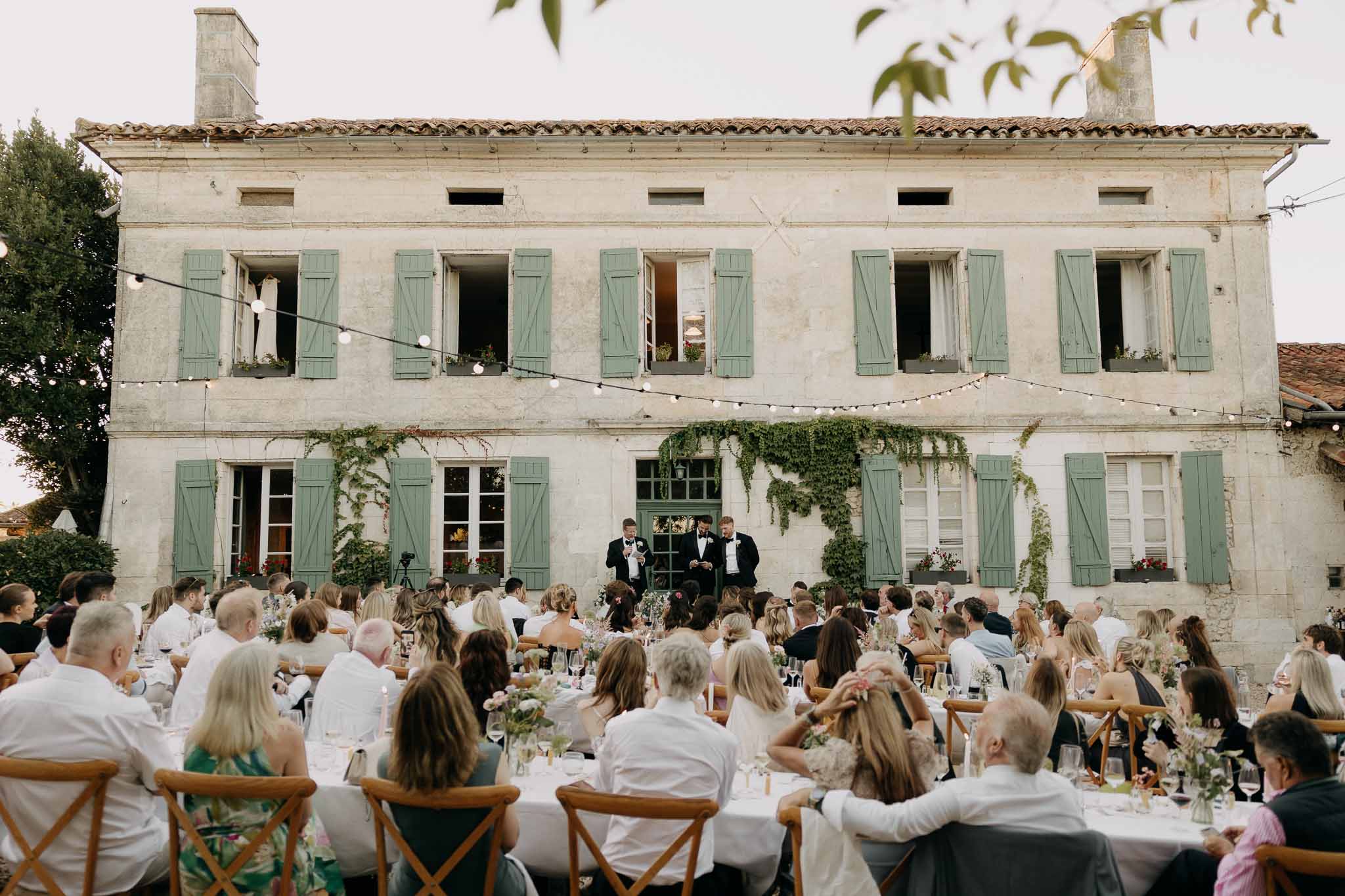Three groomsmen delivering speeches to 80 guests at festoon-lit manor with sage shutters and ivy facade