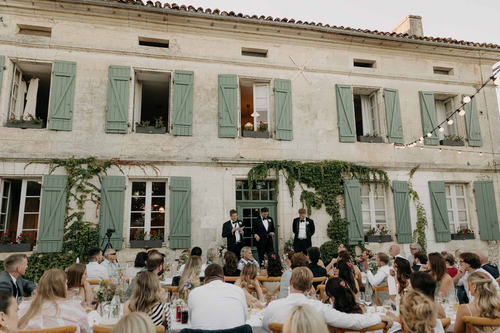 Outdoor wedding reception dinner in stone manor courtyard with guests seated at long tables and groomsmen giving speech