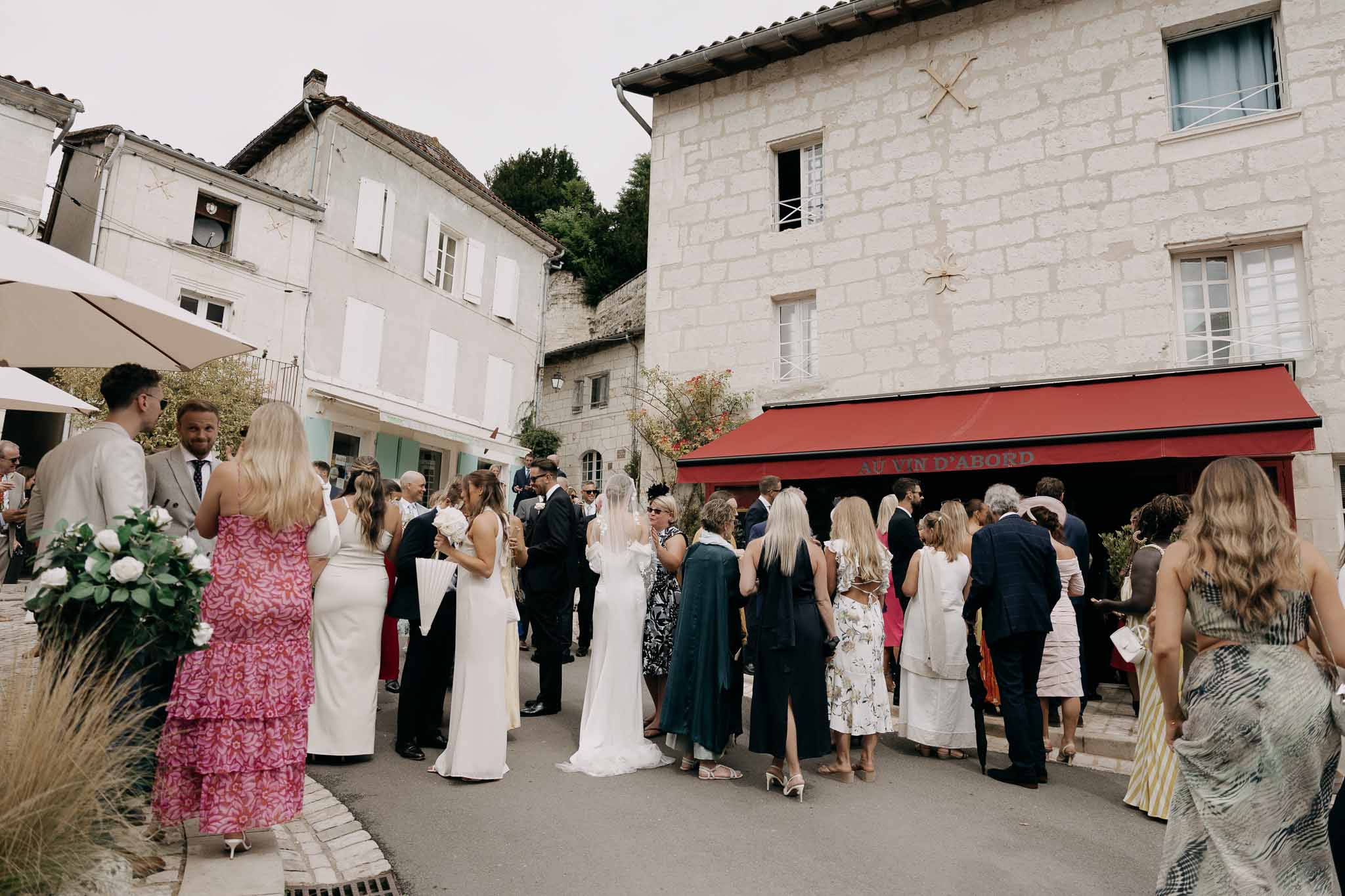 Wedding group mingling in French village square with bride in veil and bridesmaids in white before red-awning wine bar