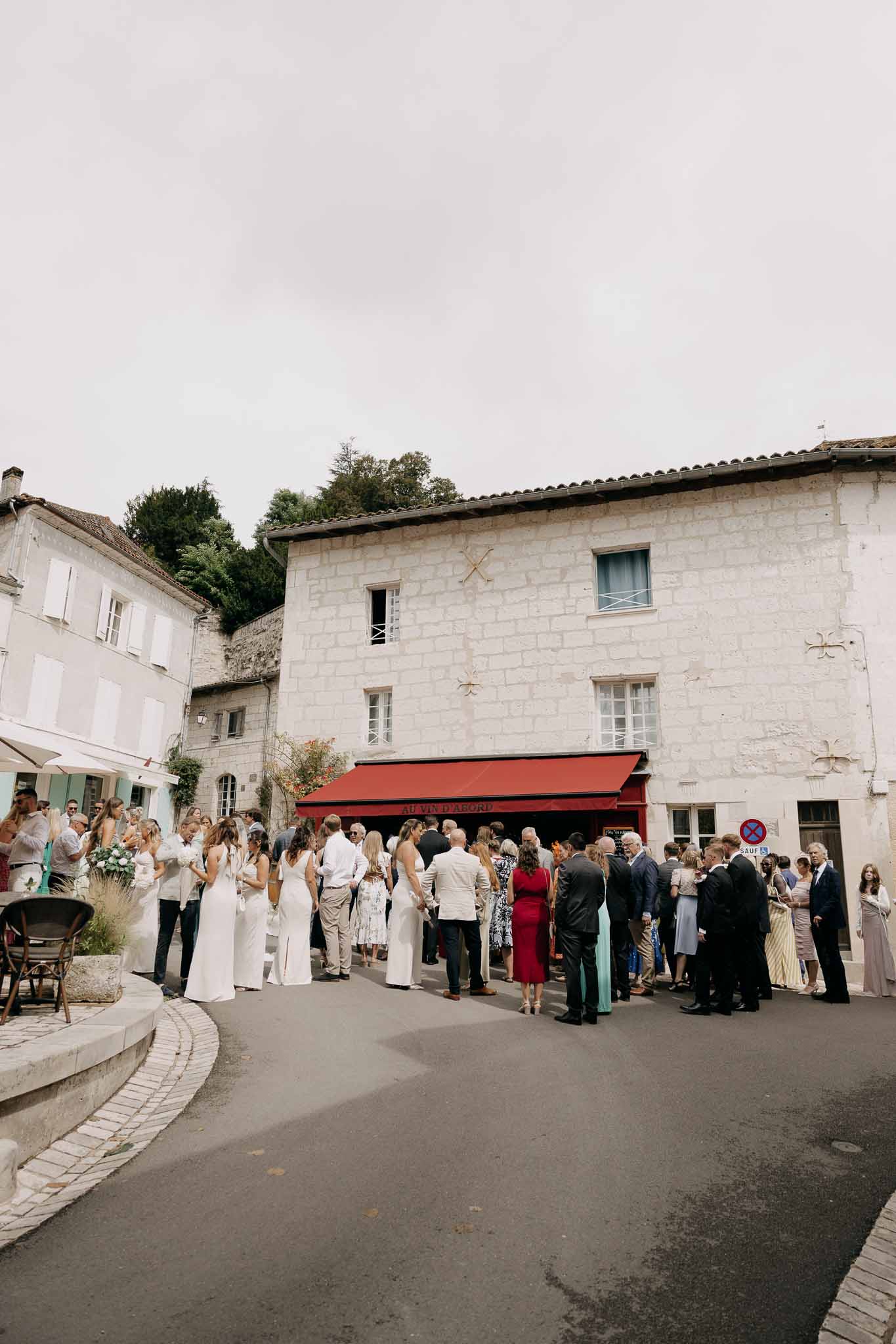 Forty guests mingling on village square before limestone building with red awning bridal party in white dresses