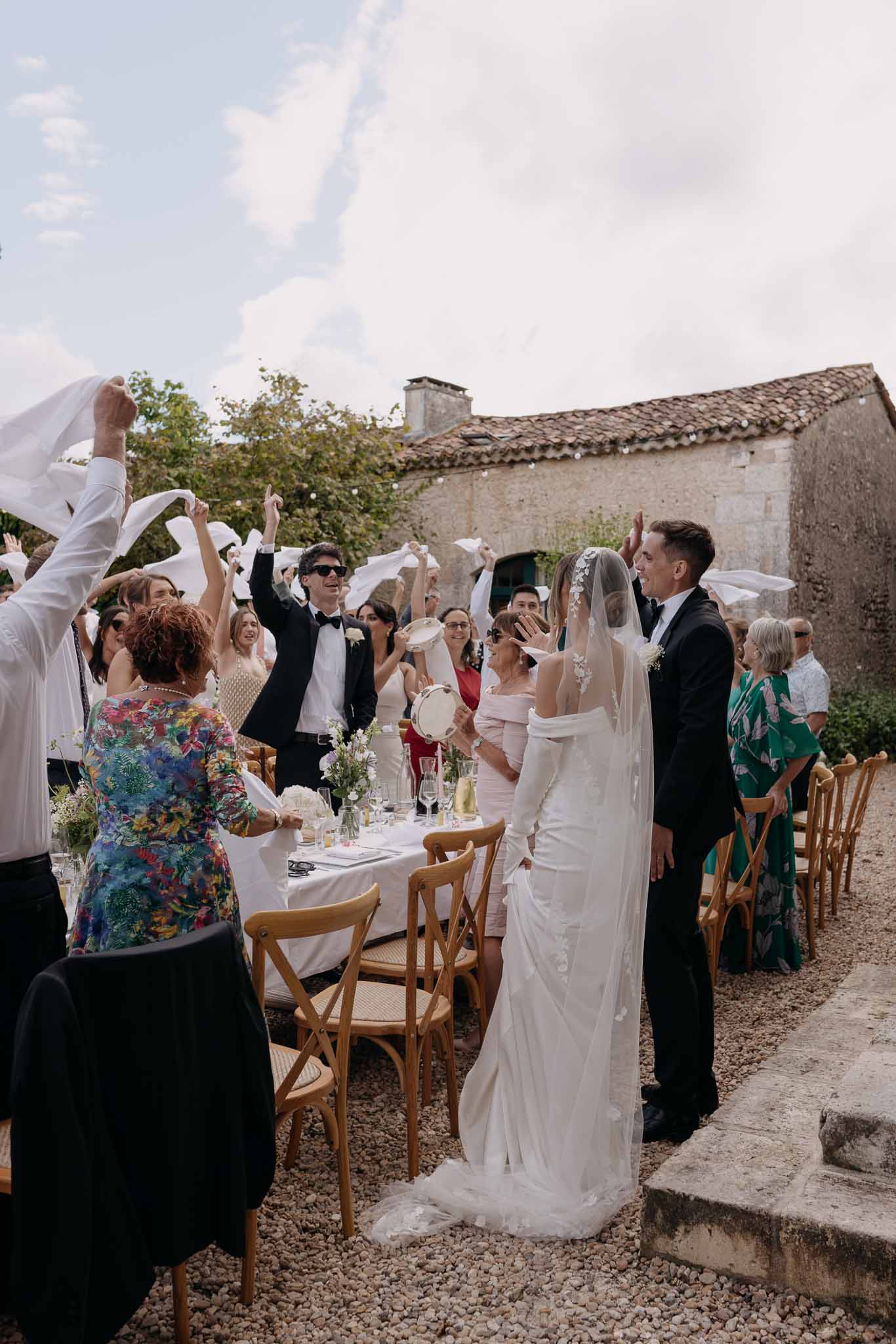 Guests waving white napkins as couple enters gravel courtyard reception with wildflower centerpieces