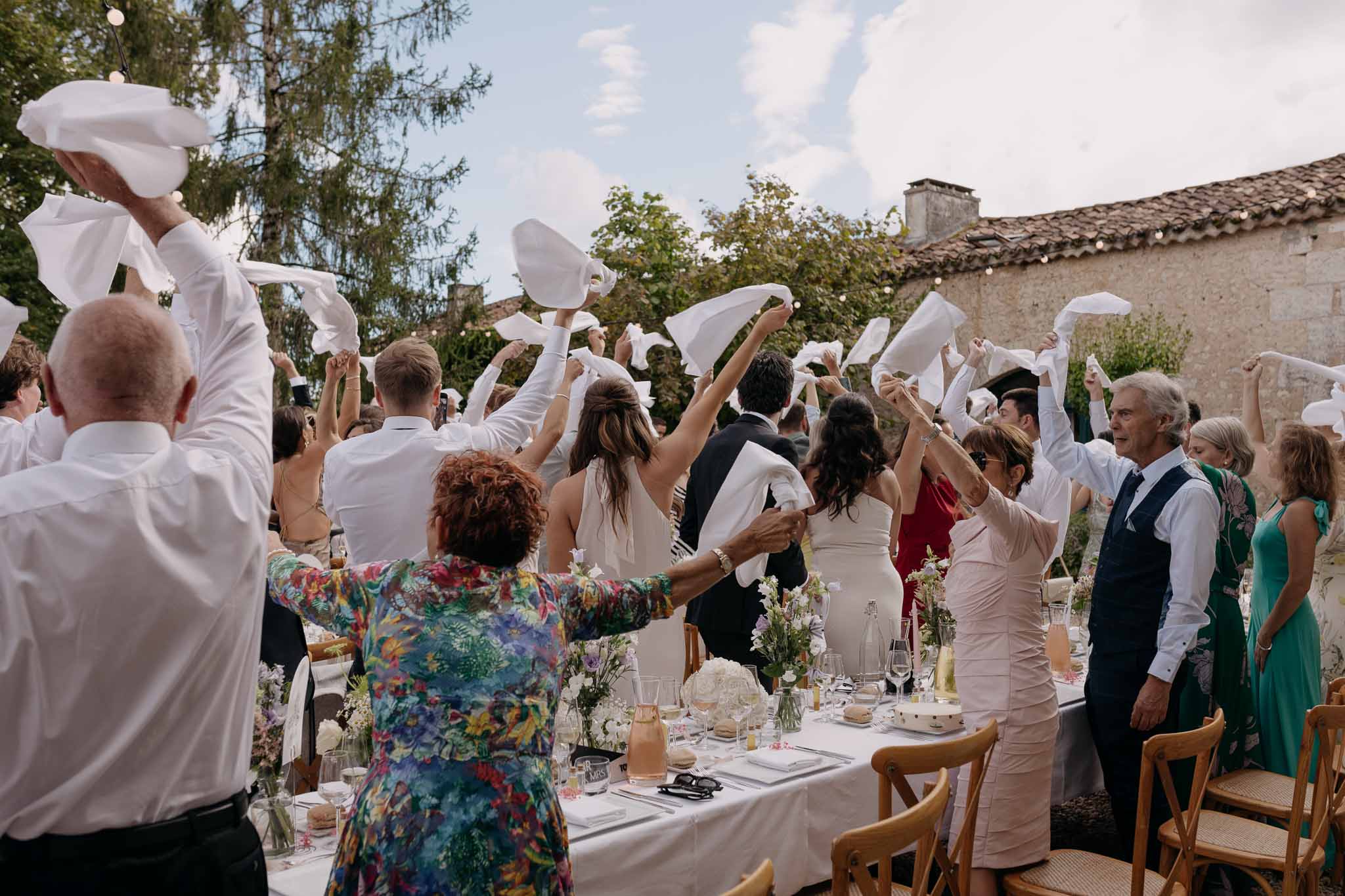 Guests wave white napkins as bride and groom stand at long tables with hydrangea centerpieces in courtyard
