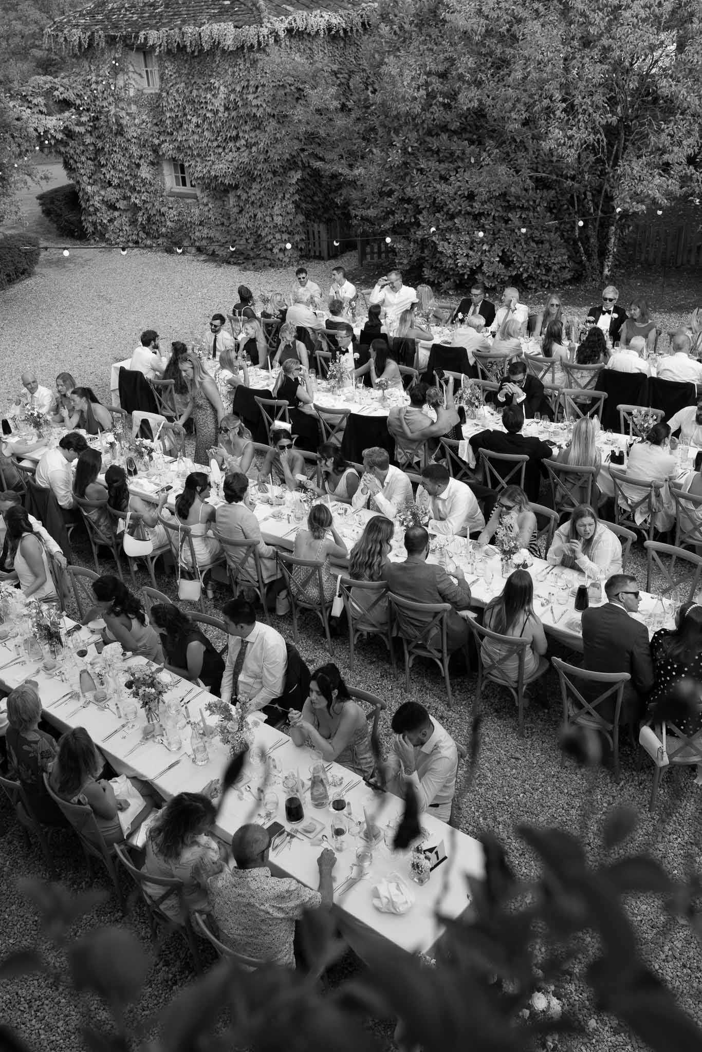 Black and white elevated view of 70 guests at long tables on gravel courtyard with globe string lights