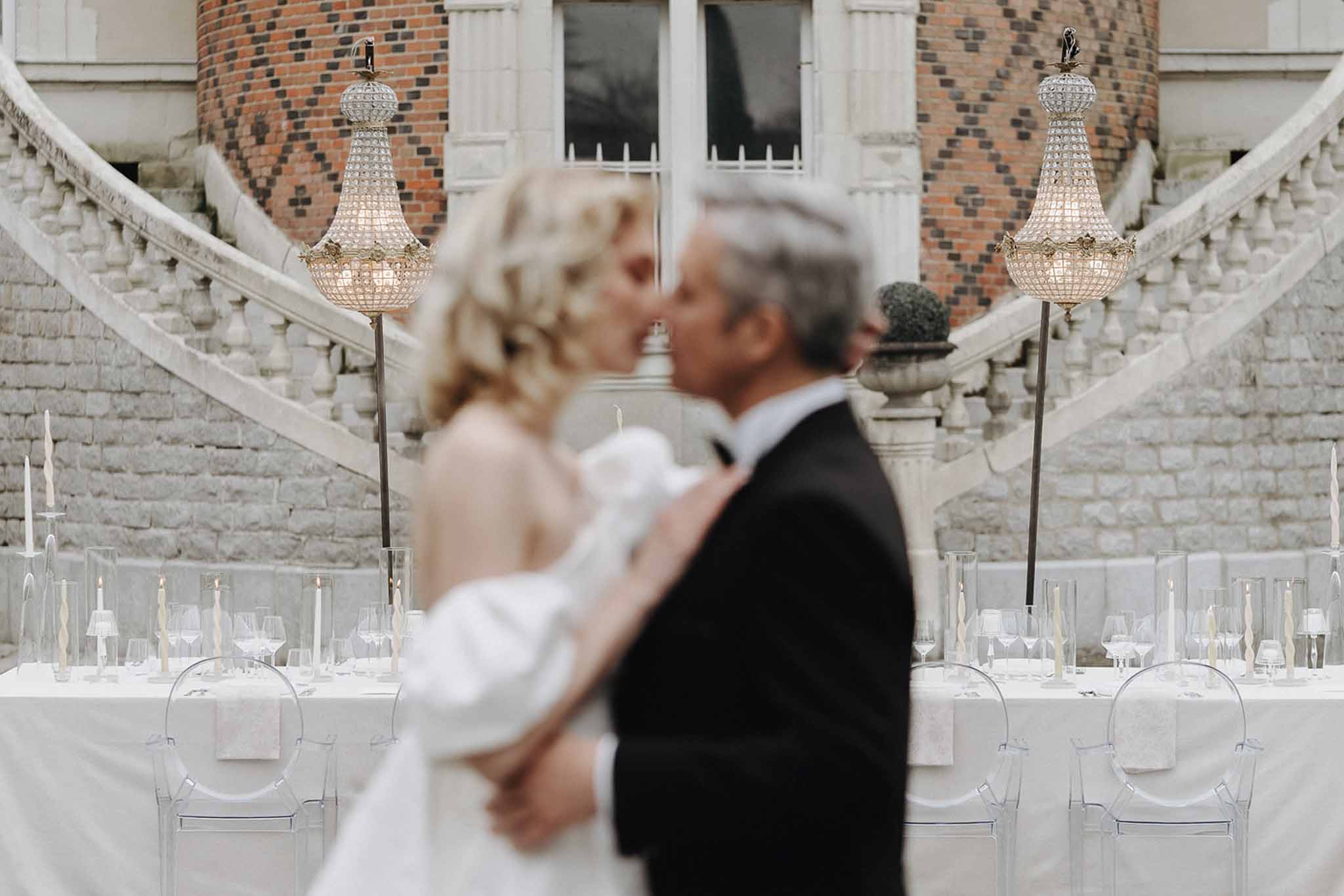 Blurred bride and groom kissing with elegant reception table and chateau double staircase in sharp focus behind
